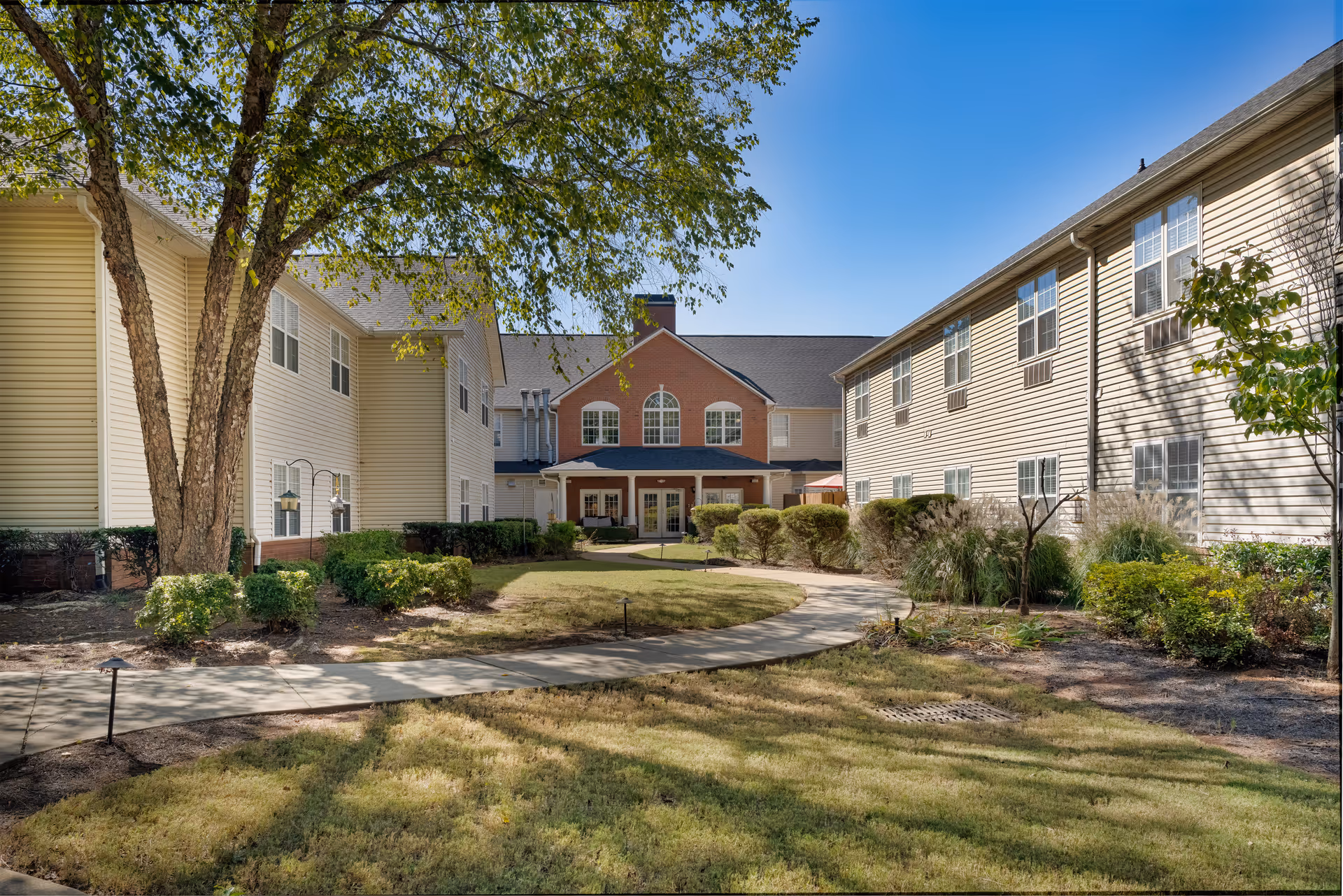 Exterior view of Azalea Gardens Mableton, showing beige buildings, a curved walkway, and landscaping.