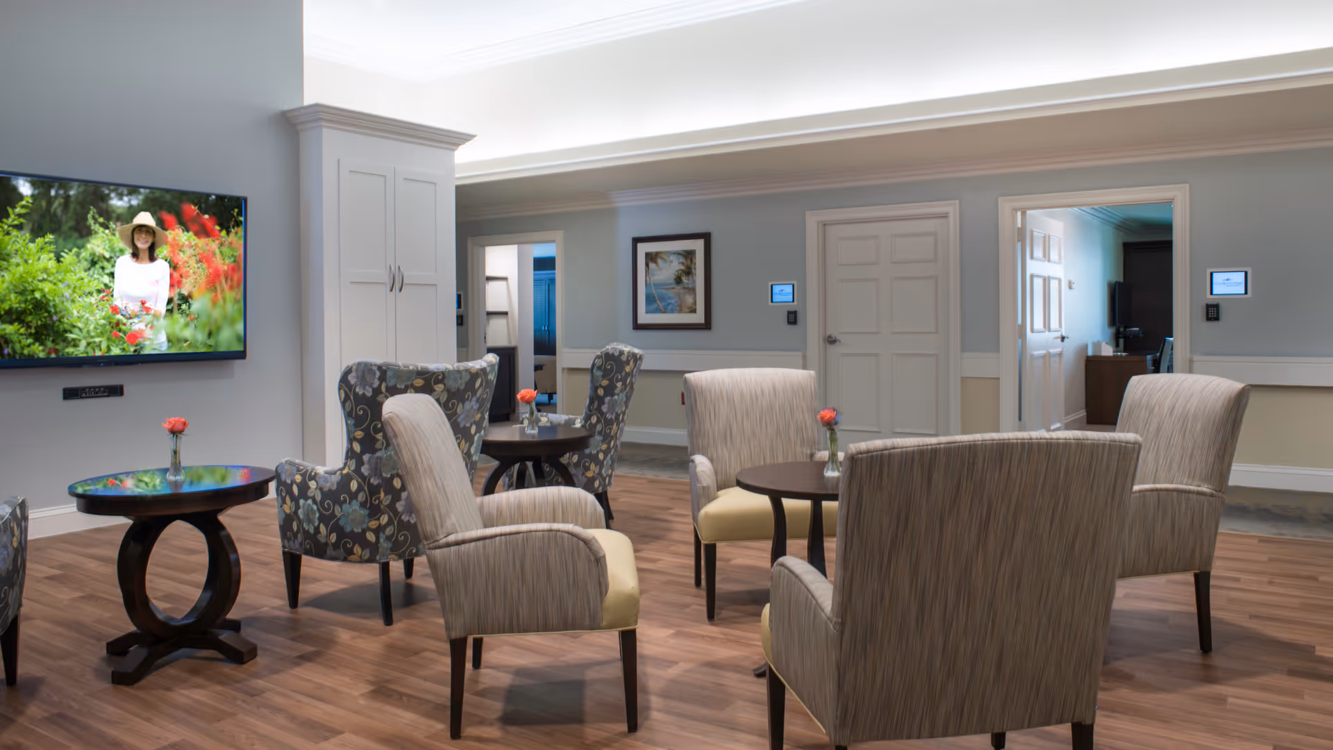 A cozy common area with several upholstered armchairs arranged around small round tables, each table holding a small vase with a single red flower. The room has wood flooring, light blue walls with white trim, and a flat-screen TV mounted on the wall displaying an image of a woman in a garden. There are doors and framed artwork visible in the background.