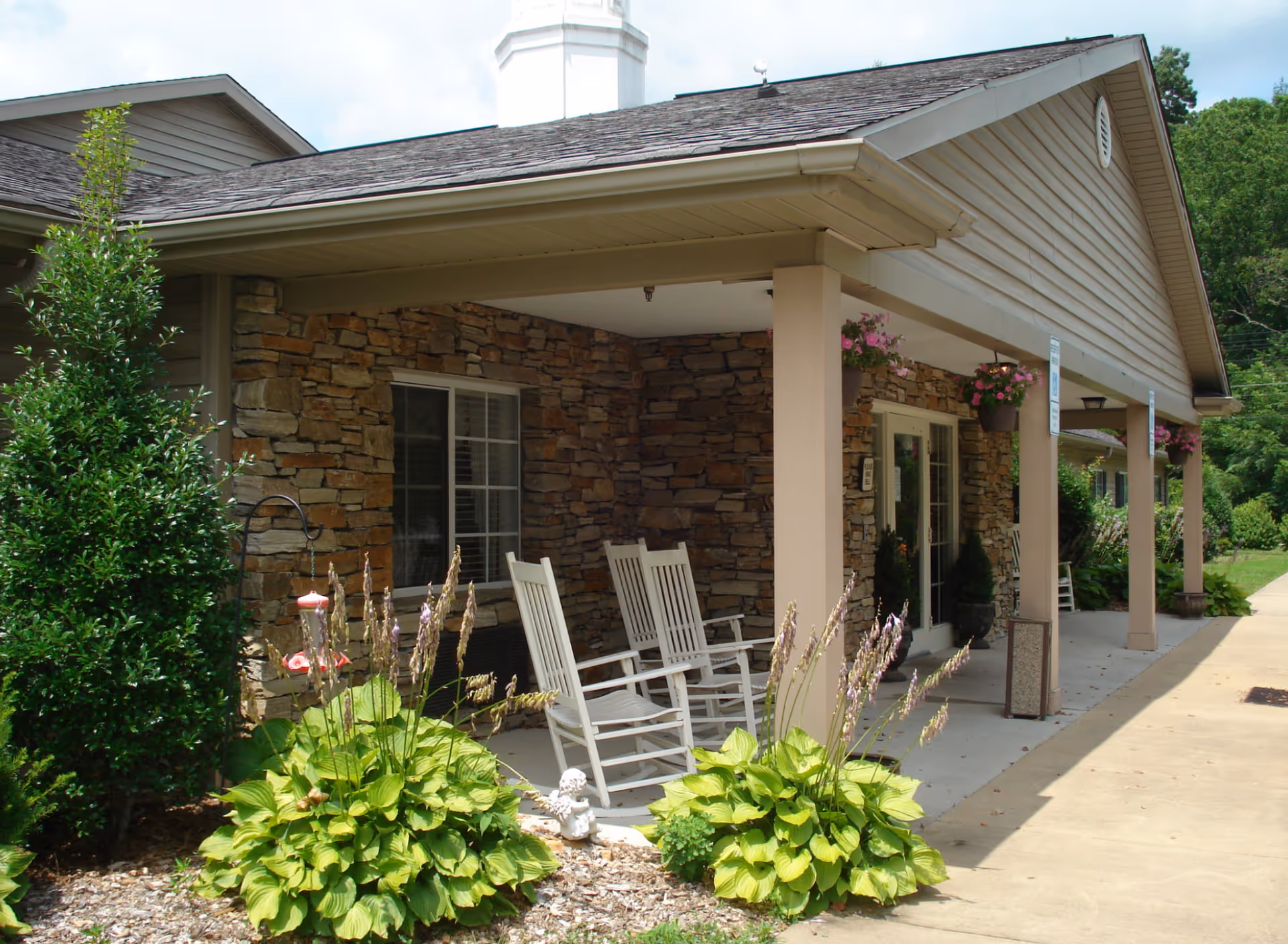 Covered porch area of a building with stone walls and beige siding, featuring white rocking chairs, hanging flower pots, and green plants in the foreground.