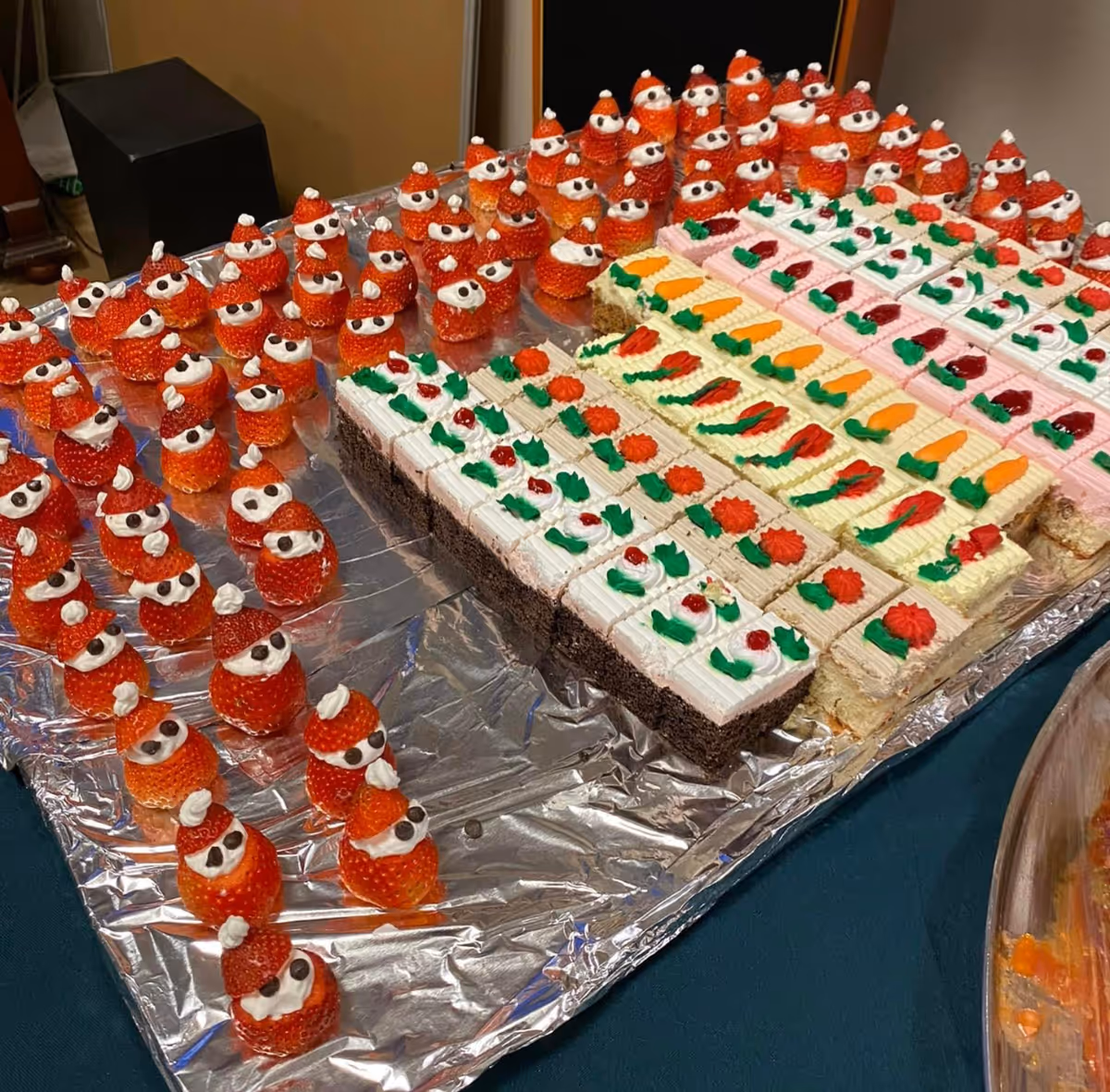 Tray of festive desserts, including strawberry 'Santa' figures and rows of decorated cake squares on foil.