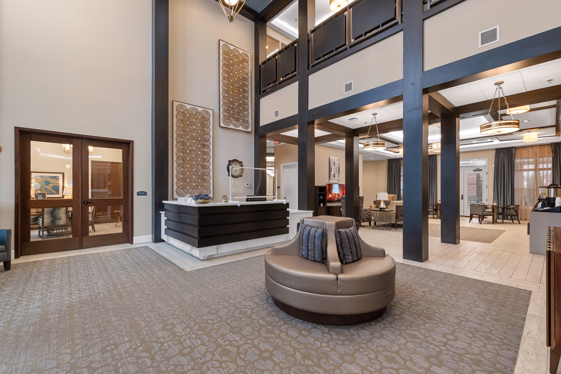 Spacious senior living facility lobby with high ceilings, a circular seating area with cushions in the center, dark wooden pillars, and a reception desk. The area features carpeted and tiled flooring, modern light fixtures, decorative wall art, and large windows with curtains allowing natural light.