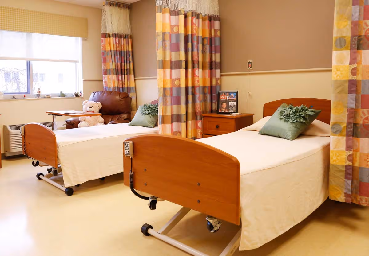 A bright and clean room with two hospital-style beds separated by colorful privacy curtains. Each bed has a green decorative pillow. There is a brown leather recliner chair with a teddy bear on it near a window with a beige valance. A wooden nightstand with framed photos is placed between the beds.