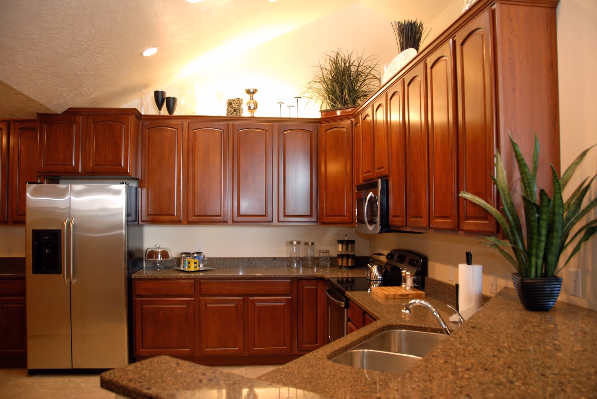 A modern kitchen with dark wooden cabinets, a stainless steel refrigerator, a built-in microwave, an electric stove, and a double sink. The countertops are granite, and there are decorative plants and kitchen items placed on the counters and above the cabinets.
