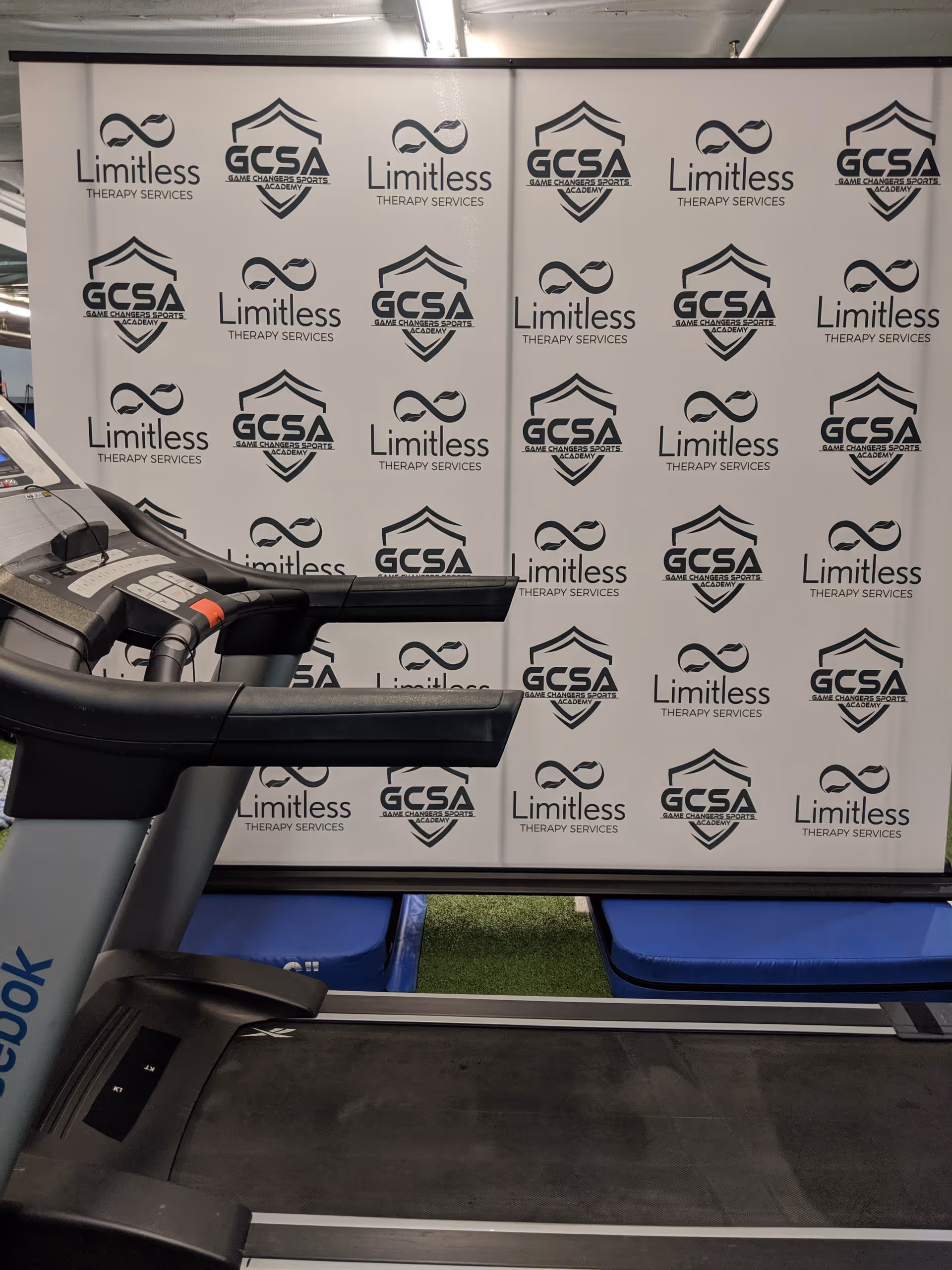 Treadmill in a gym beside a wall covered with Limitless Therapy Services and GCSA logos.
