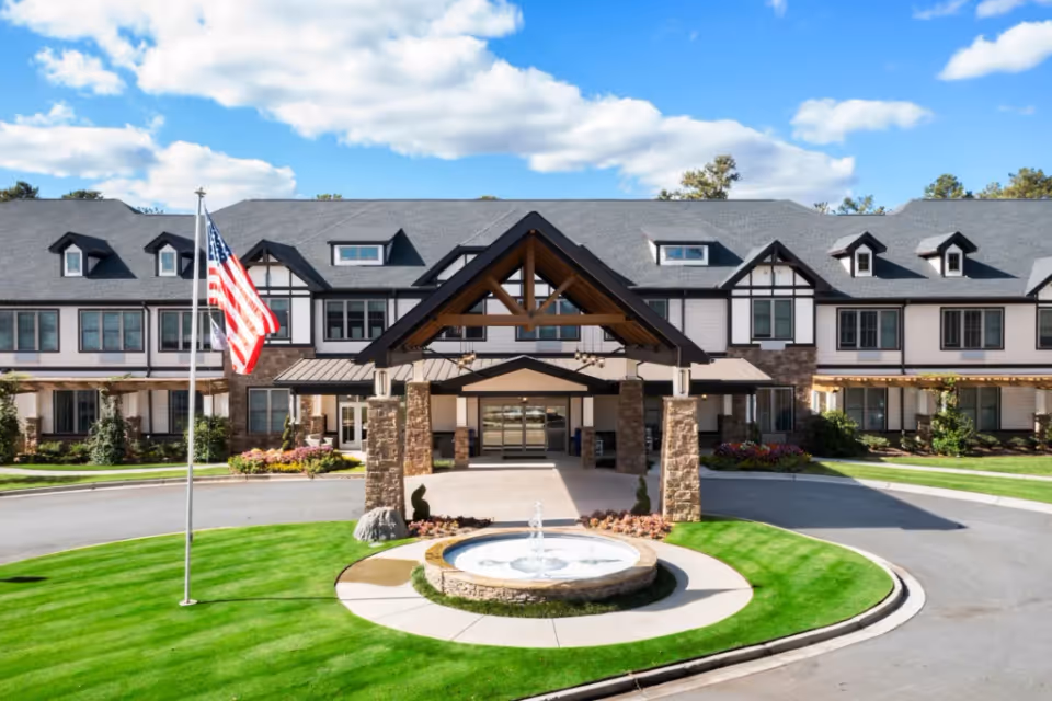 Front entrance of the Oaks at Douglasville building with a covered porte-cochere, circular fountain, and an American flag on a sunny day.