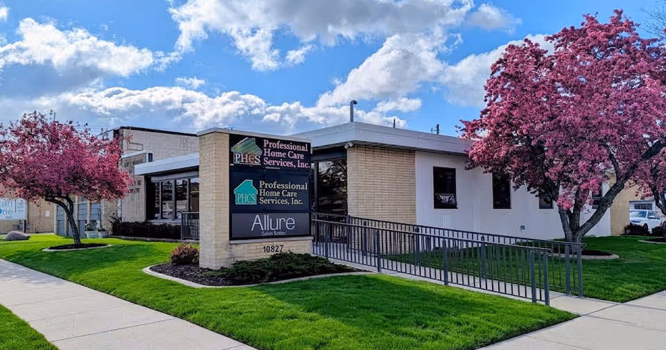 Exterior view of a senior care facility with signage, ramps, and pink flowering trees.