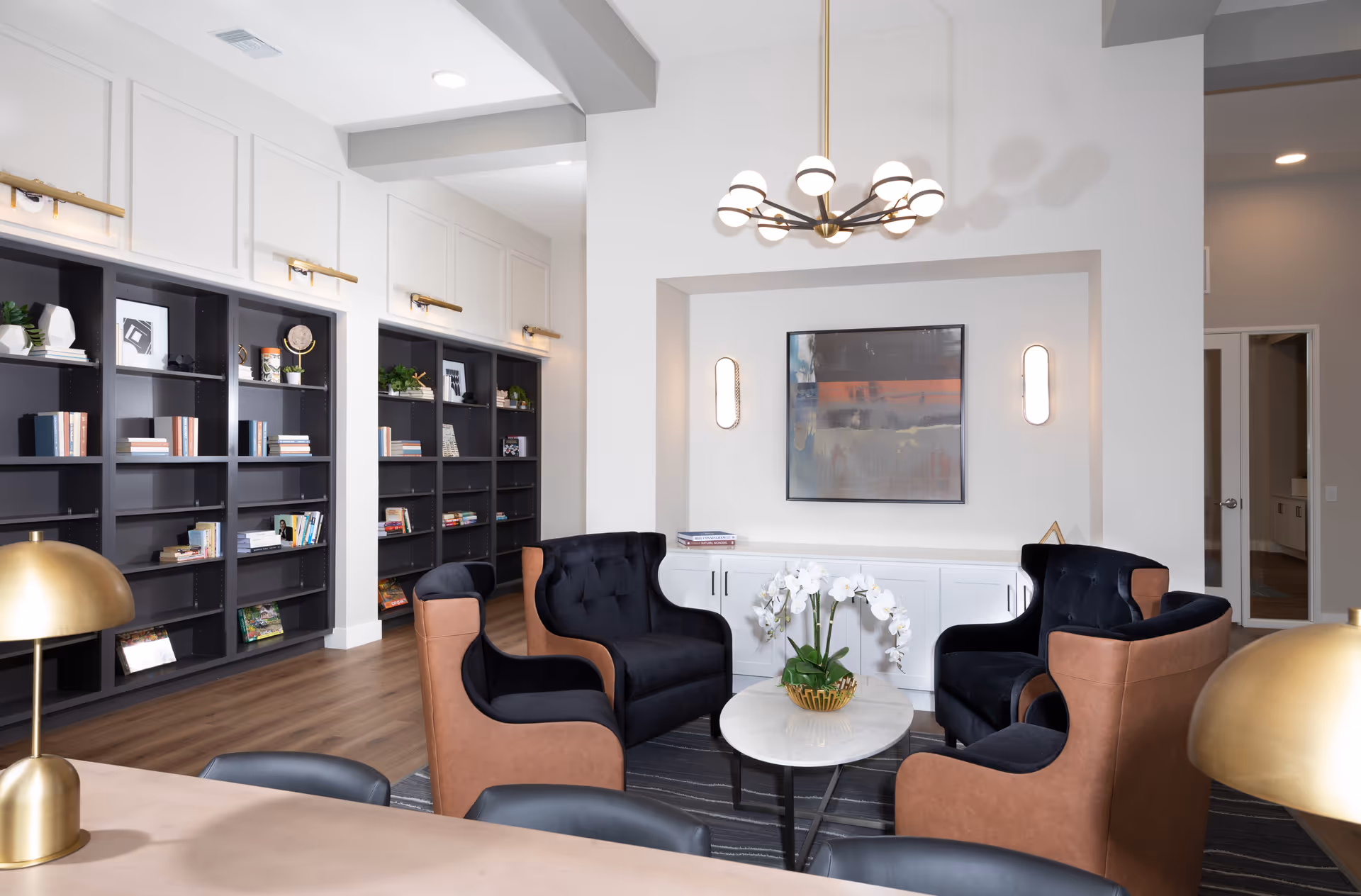 A modern living room area with four black and brown armchairs arranged around a small round white marble table with a white orchid centerpiece. Behind the seating area is a white cabinet with a colorful abstract painting and two wall sconces. To the left, there are tall black bookshelves filled with books and decorative items. The room has wooden flooring, a contemporary chandelier, and gold table lamps in the foreground.