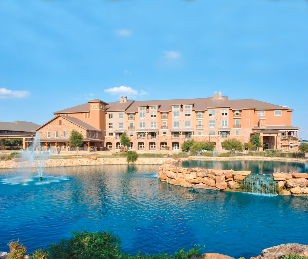A large multi-story building with a stone and stucco exterior situated behind a pond with a small waterfall and a fountain, under a clear blue sky.