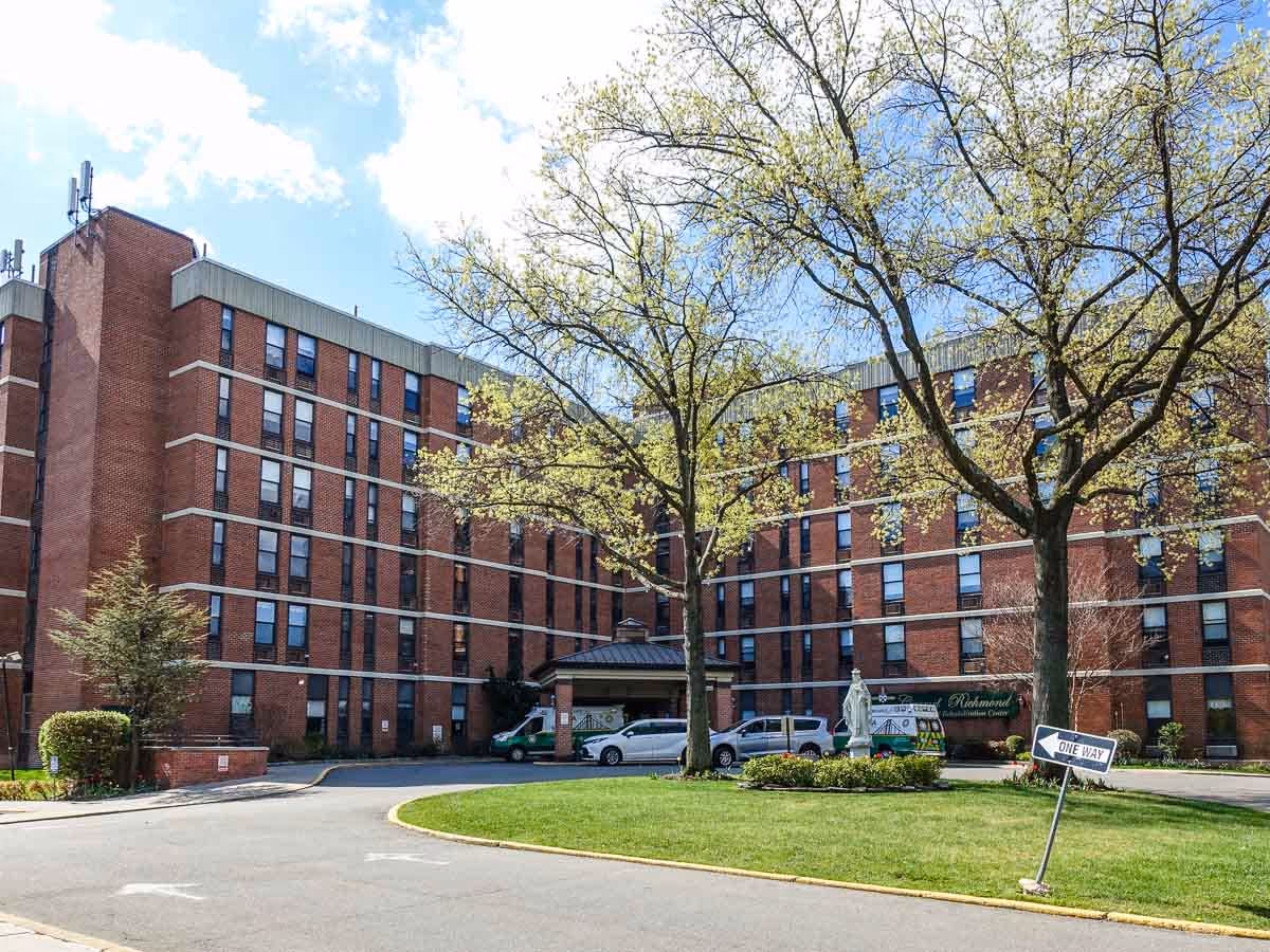 Large brick senior living building with trees, vehicles, and a curved entry driveway.