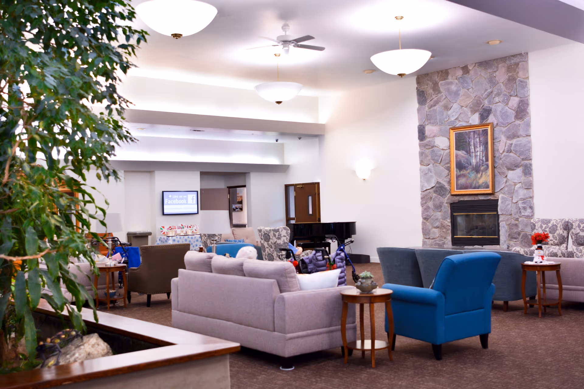 Bright communal lounge with sofas and armchairs arranged around a stone fireplace and a piano, with plants and ceiling lights.