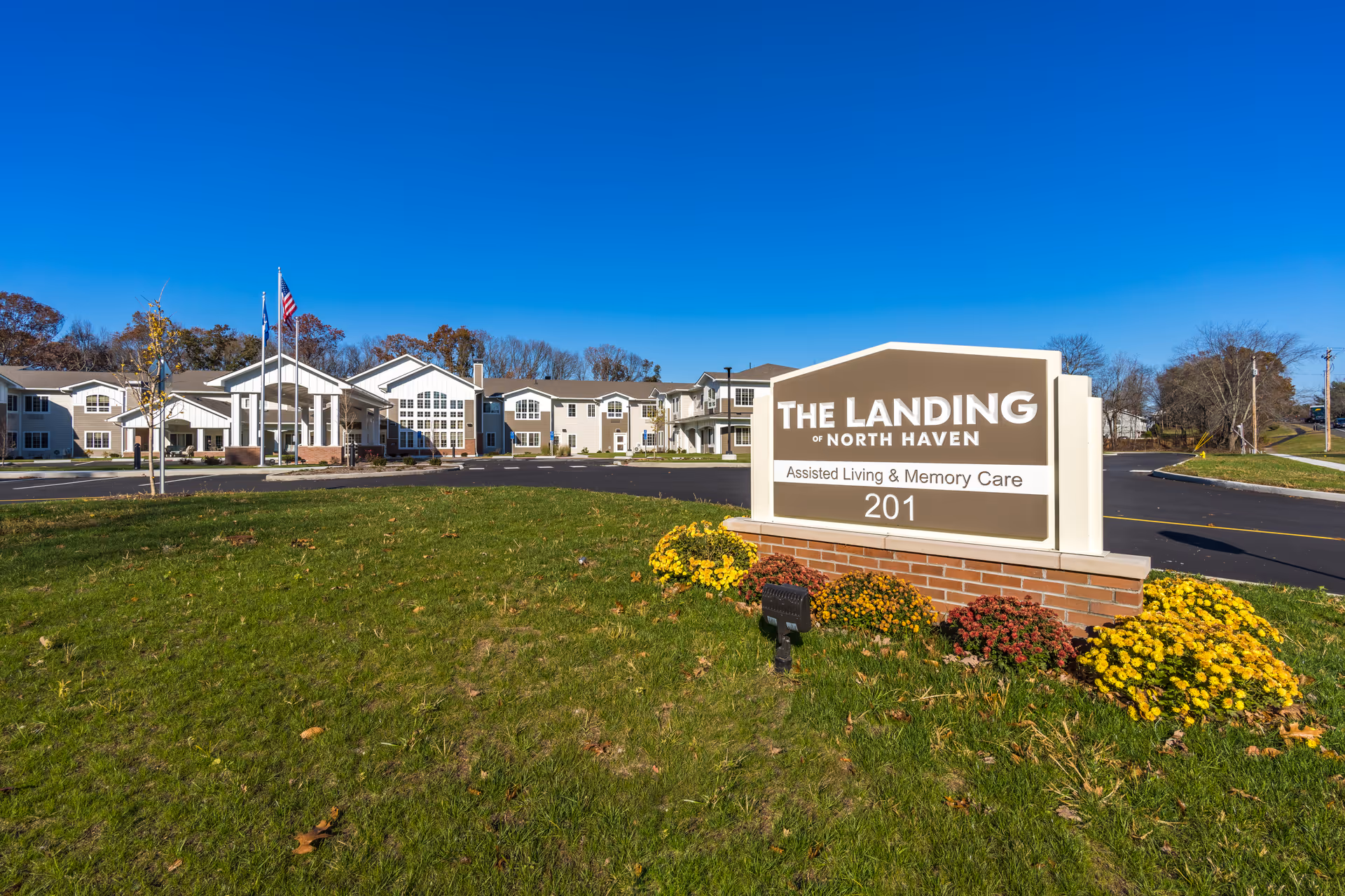 Exterior view of The Landing of North Haven assisted living and memory care facility on a clear sunny day, showing the building, entrance with flags, and a sign with the facility name and address 201 surrounded by flowers and green grass.