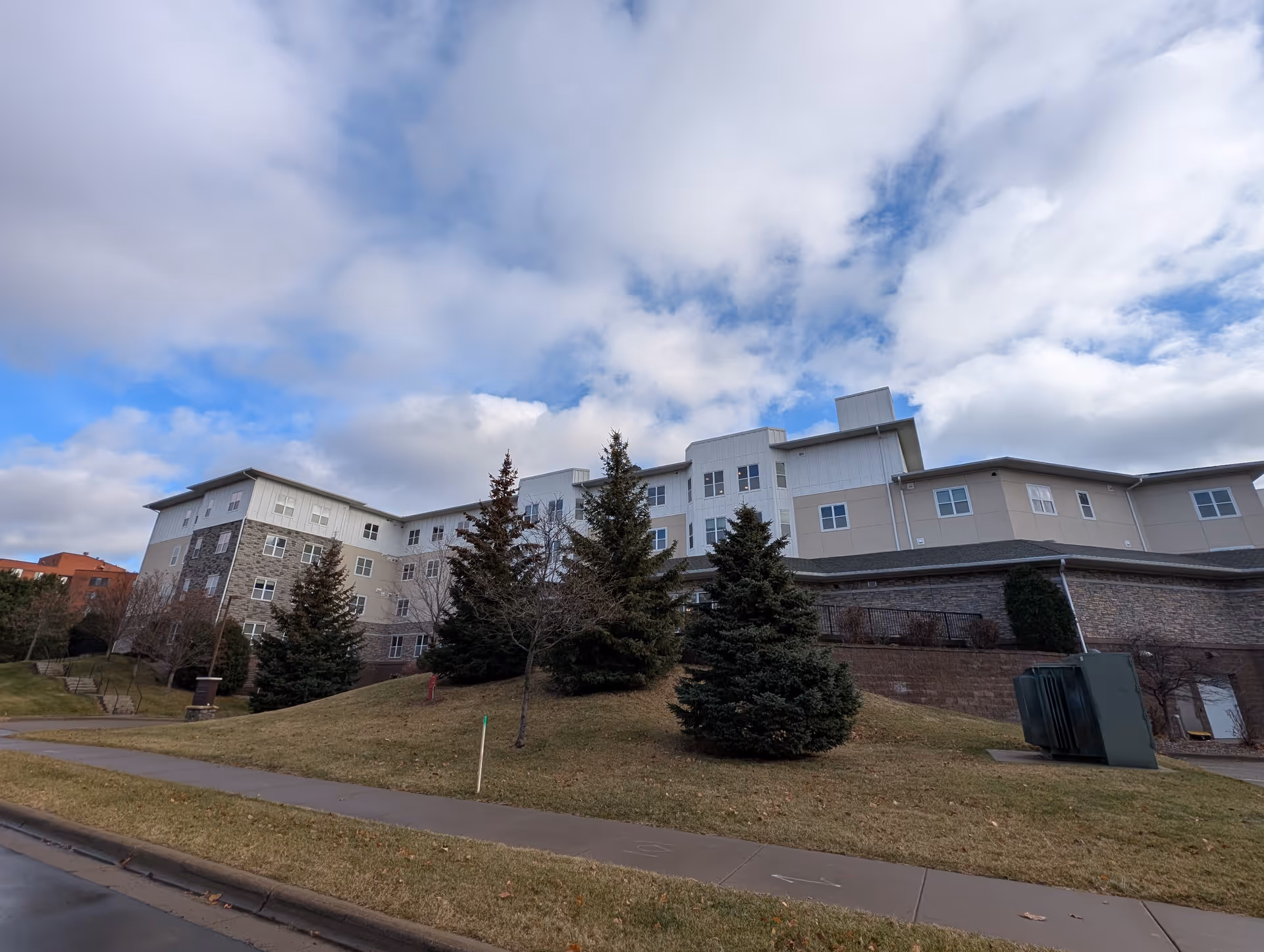 Multi-story residential building with stone and light-colored siding behind evergreen trees on a grassy slope under a cloudy blue sky.