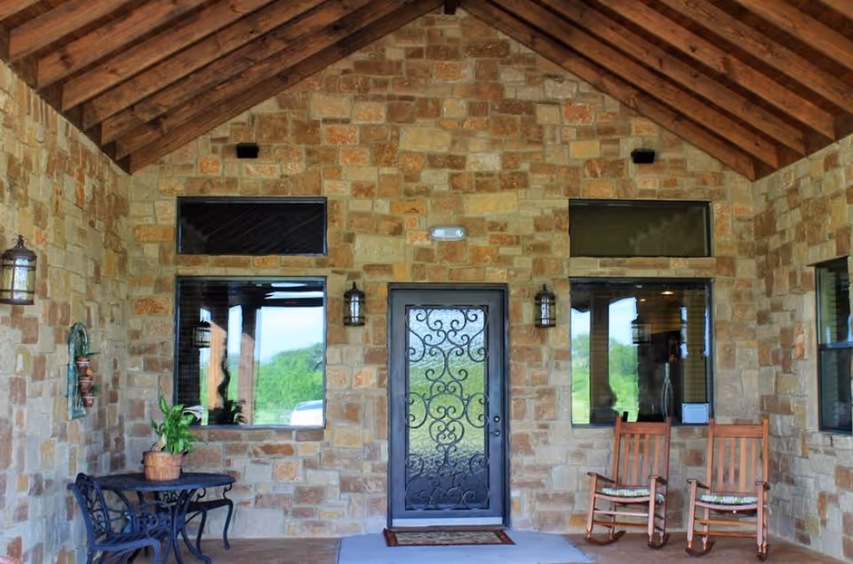 Stone-faced front porch with decorative iron door, two rocking chairs, and potted plants.