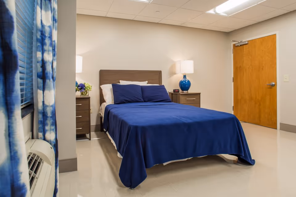 A neat bedroom with a bed dressed in blue linens, two bedside tables with lamps, blue patterned curtains and a wooden door.