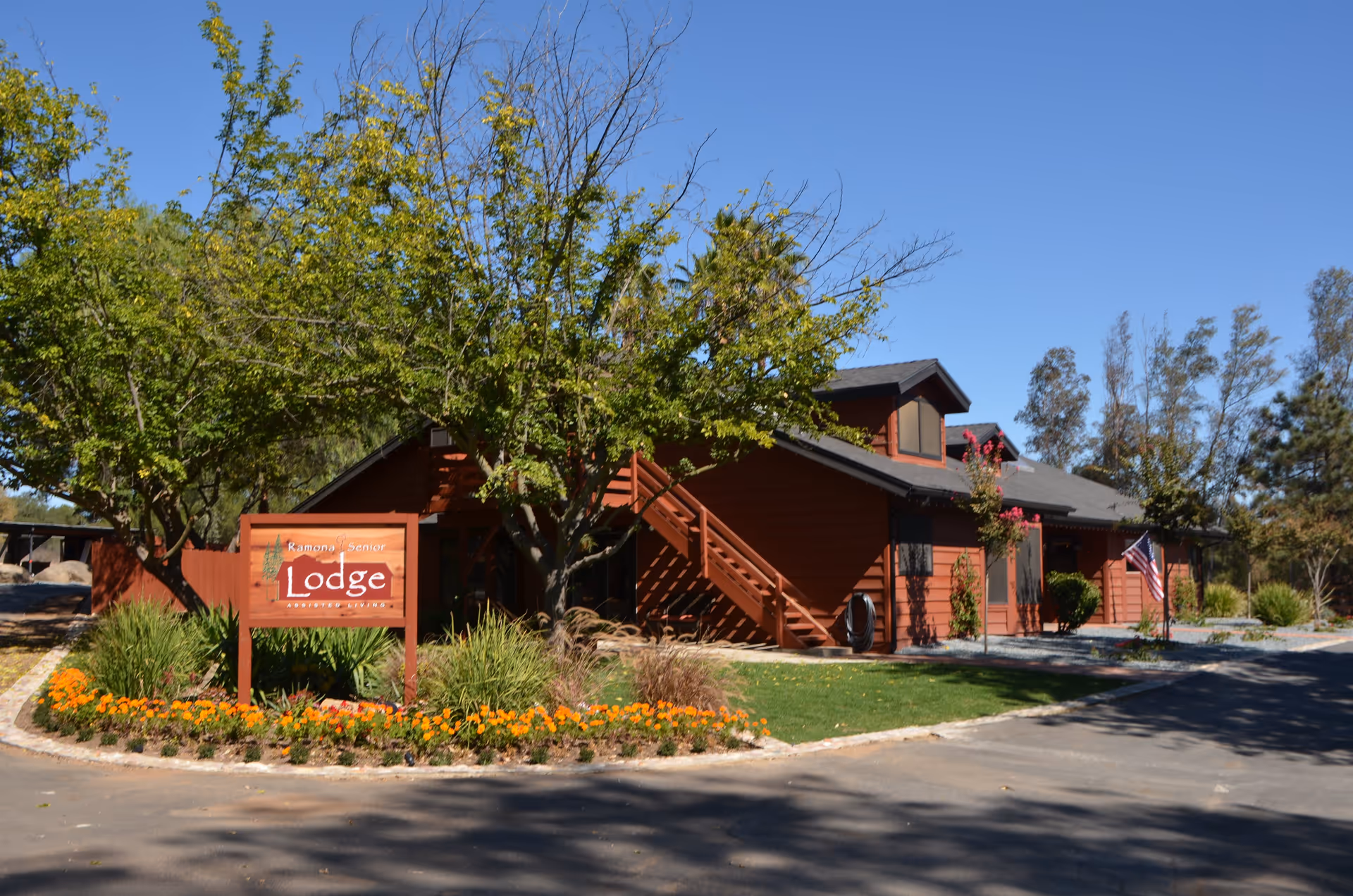 Front exterior of a red two-story lodge building with a 'Lodge' sign, trees, and landscaped flowerbeds under a clear blue sky.