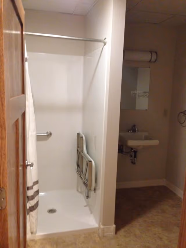 A small bathroom area with a walk-in shower featuring a white shower curtain and a foldable shower seat attached to the wall. There is a grab bar inside the shower for support. Adjacent to the shower is a small sink with a mirror above it and a towel ring on the wall. The floor has a beige patterned linoleum surface.