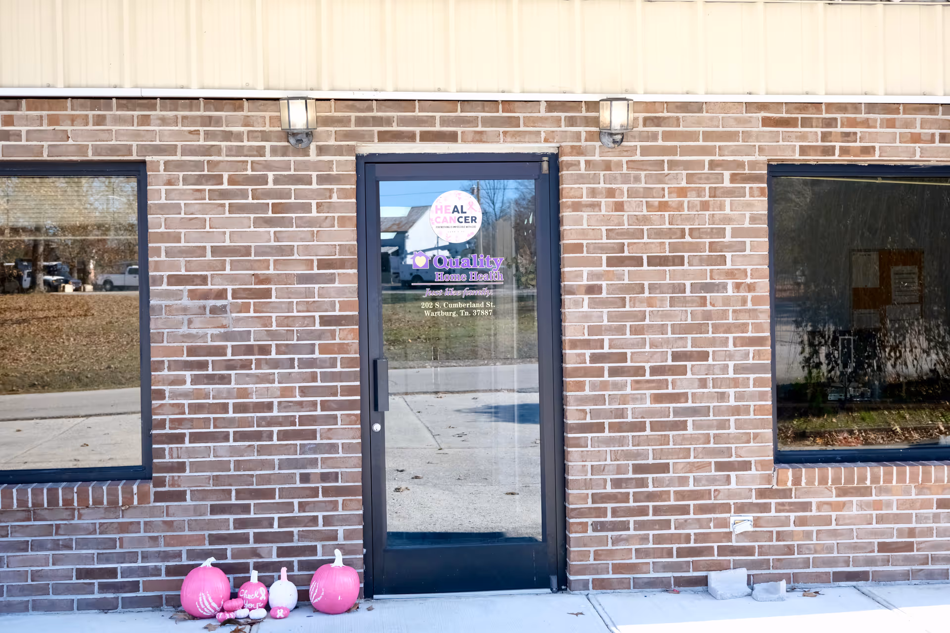 Brick building exterior with glass entrance door, sign reading Quality Home Health, pumpkins outside the entrance.