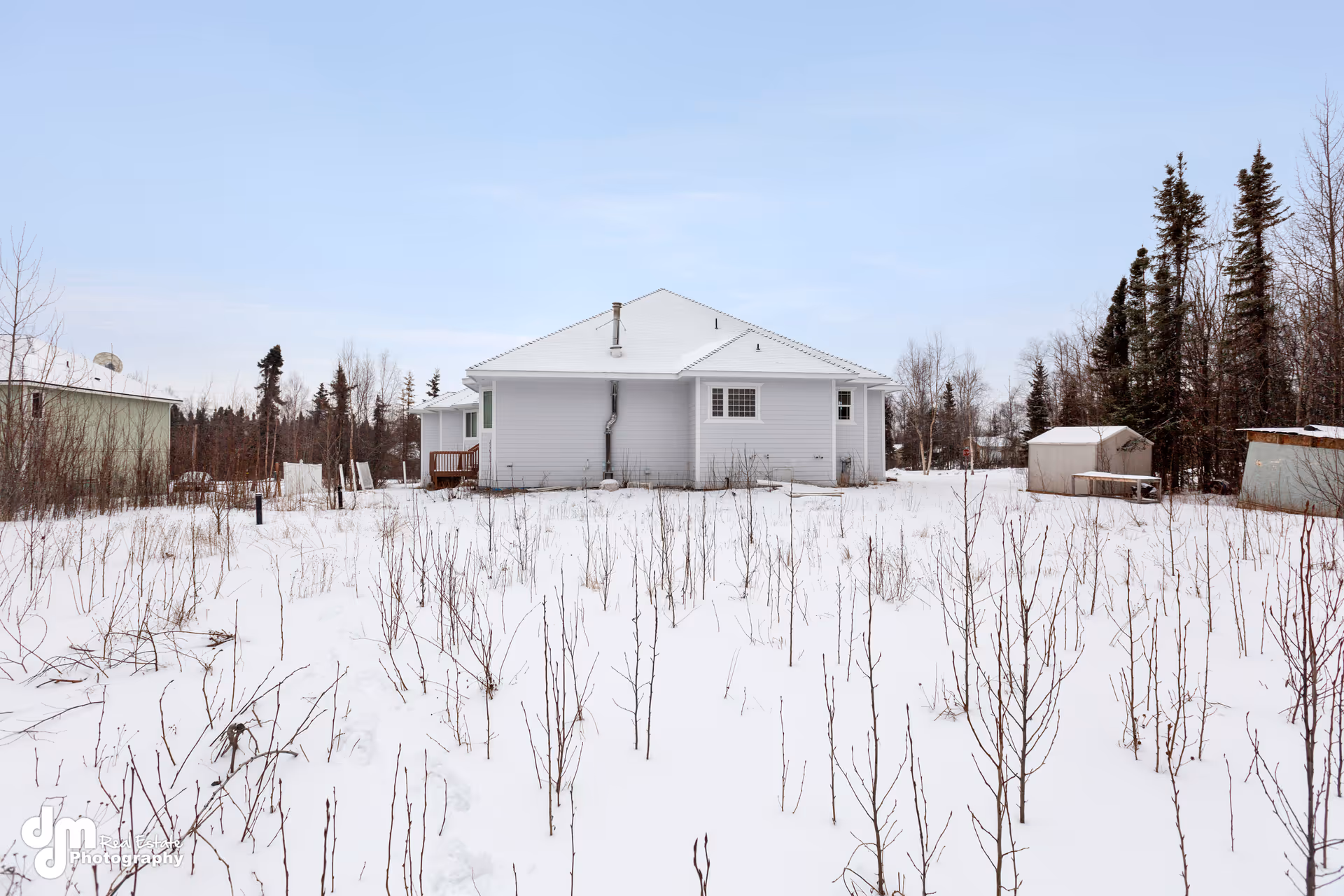 Snow-covered, single-story white house exterior with a small deck amid leafless trees.