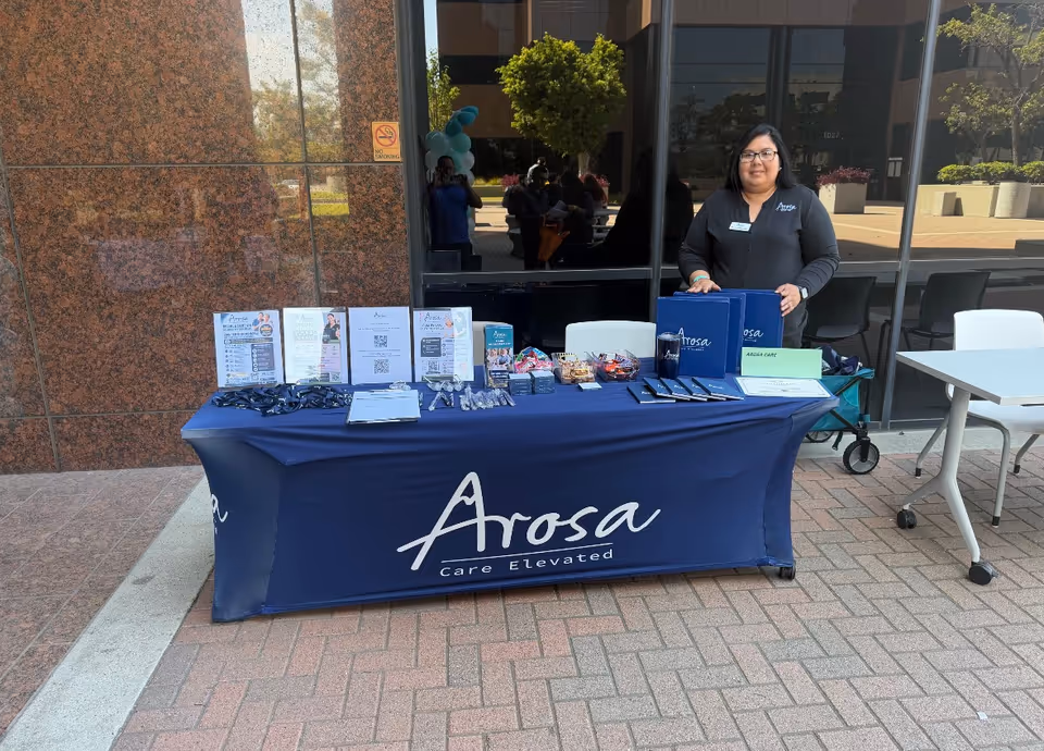 Staff member at a blue Arosa table with brochures outdoors in front of a modern building.