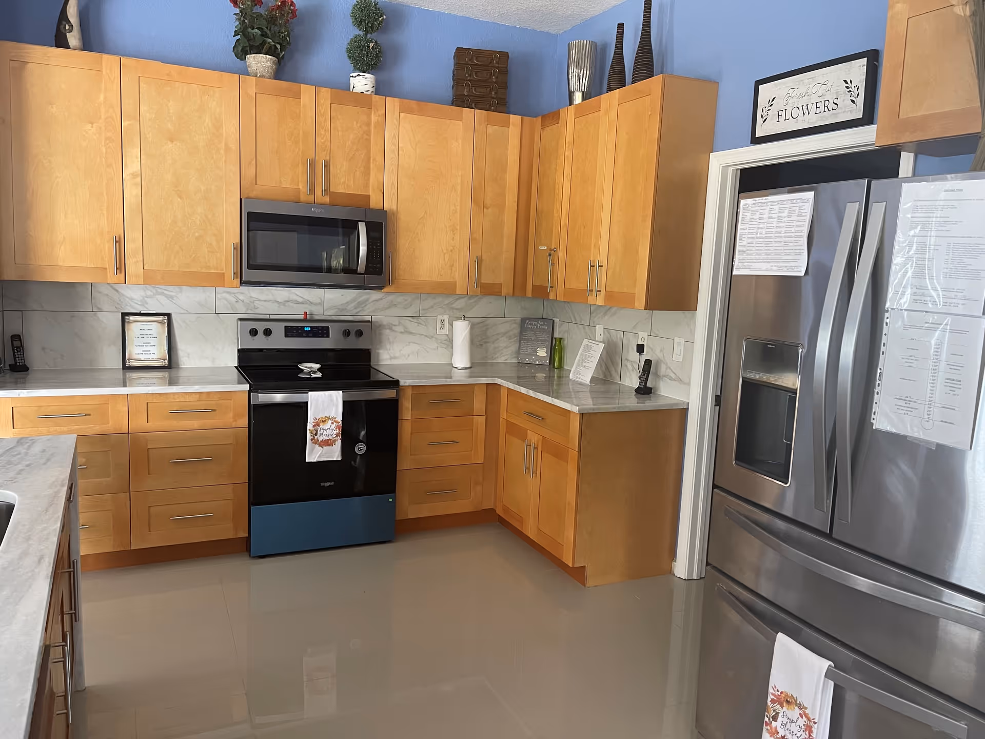 Bright modern kitchen with light wood cabinets, stainless steel refrigerator and stove, microwave, marble countertops, and blue-painted walls.