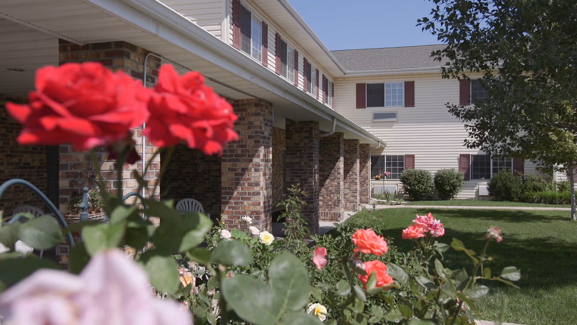 Exterior view of a senior living facility building with brick and siding walls, multiple windows with red shutters, a covered walkway supported by brick columns, and a garden with blooming red, pink, and white roses in the foreground under a clear sky.
