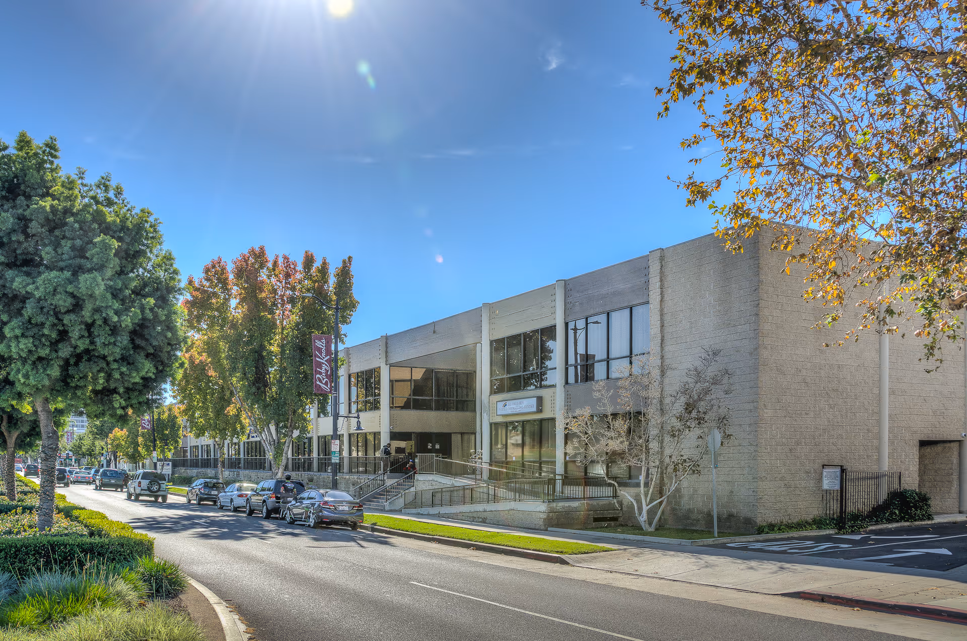 Exterior view of a modern senior facility front with a street, trees, and parked cars.