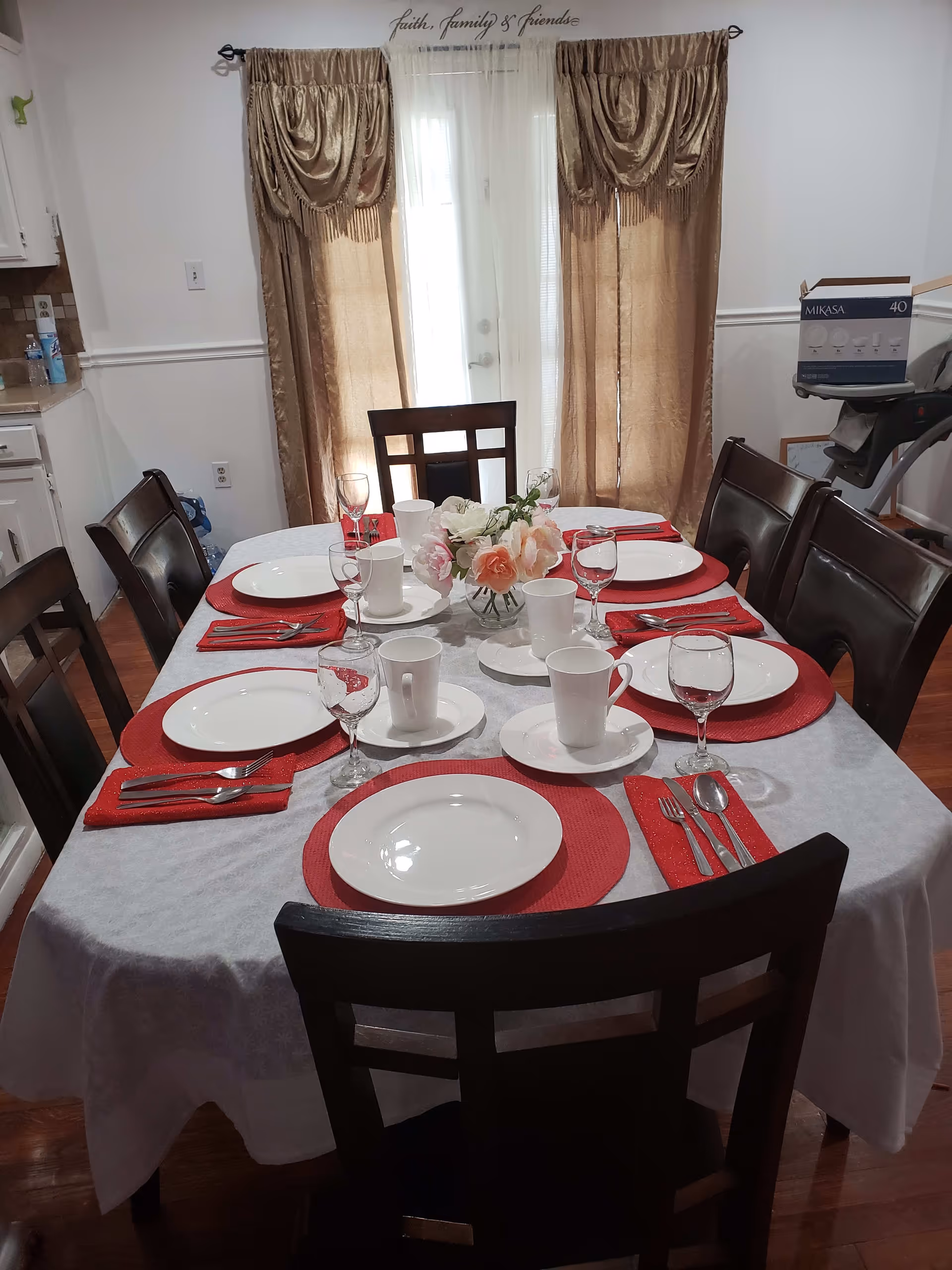 A dining table set for six people with white plates, cups, wine glasses, and silverware on red placemats and napkins. A floral centerpiece is in the middle of the table. Behind the table are beige curtains covering a glass door, with the words 'faith, family & friends' written above.