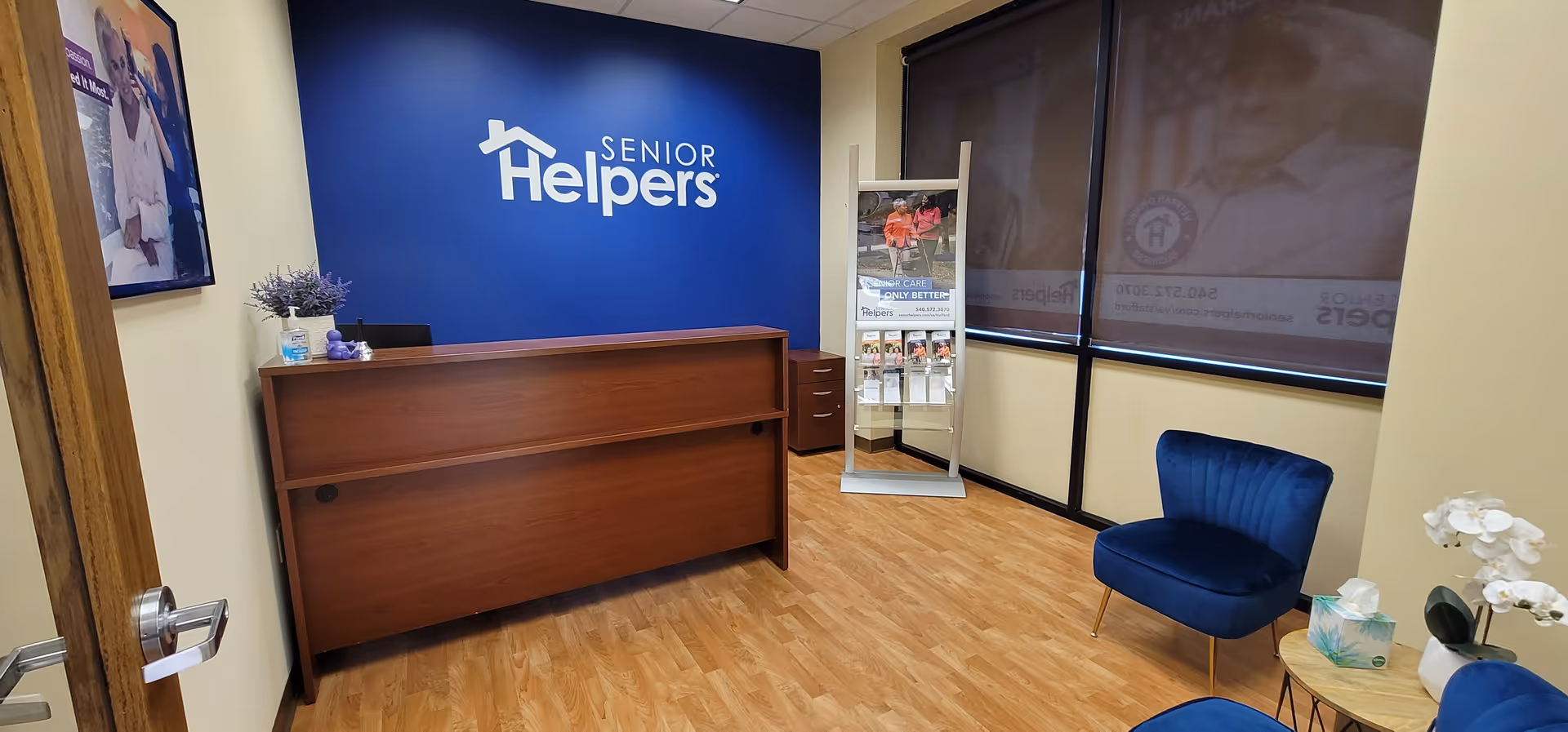 Reception area featuring a wooden desk, blue Senior Helpers logo wall, brochure rack, and blue seating.