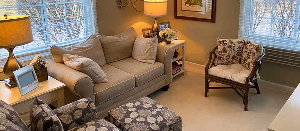 A cozy living room with a beige couch, floral-patterned armchair and ottoman, side tables with lamps, framed art, and windows with blinds.