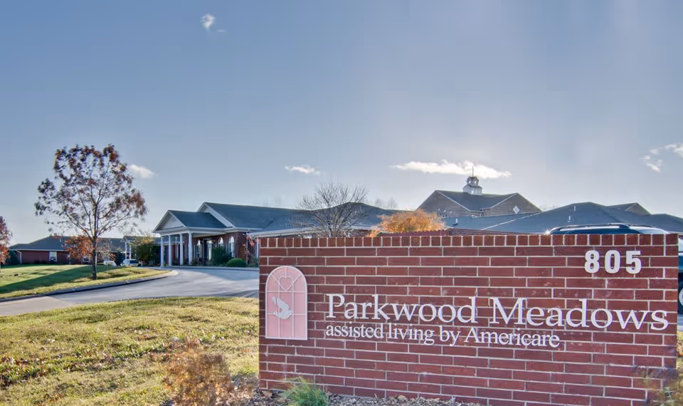 Exterior view of Parkwood Meadows Senior Living facility with a brick sign in the foreground displaying the name and address number 805. The building has a pitched roof and a covered entrance with columns. Trees and a clear blue sky are visible in the background.