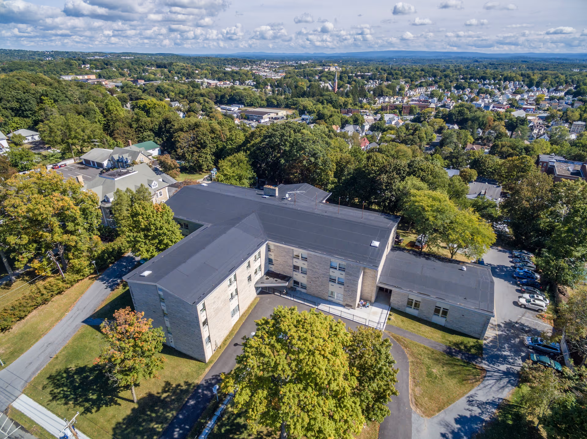 Aerial view of Highland Rehabilitation & Nursing Center, a multi-story building surrounded by trees and greenery, with a parking area and a residential neighborhood in the background under a partly cloudy sky.