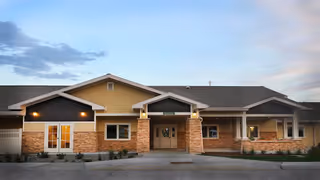 Exterior view of a single-story building with a combination of yellow siding and brick facade, featuring a central entrance with a covered porch and multiple windows, under a partly cloudy sky at dusk.