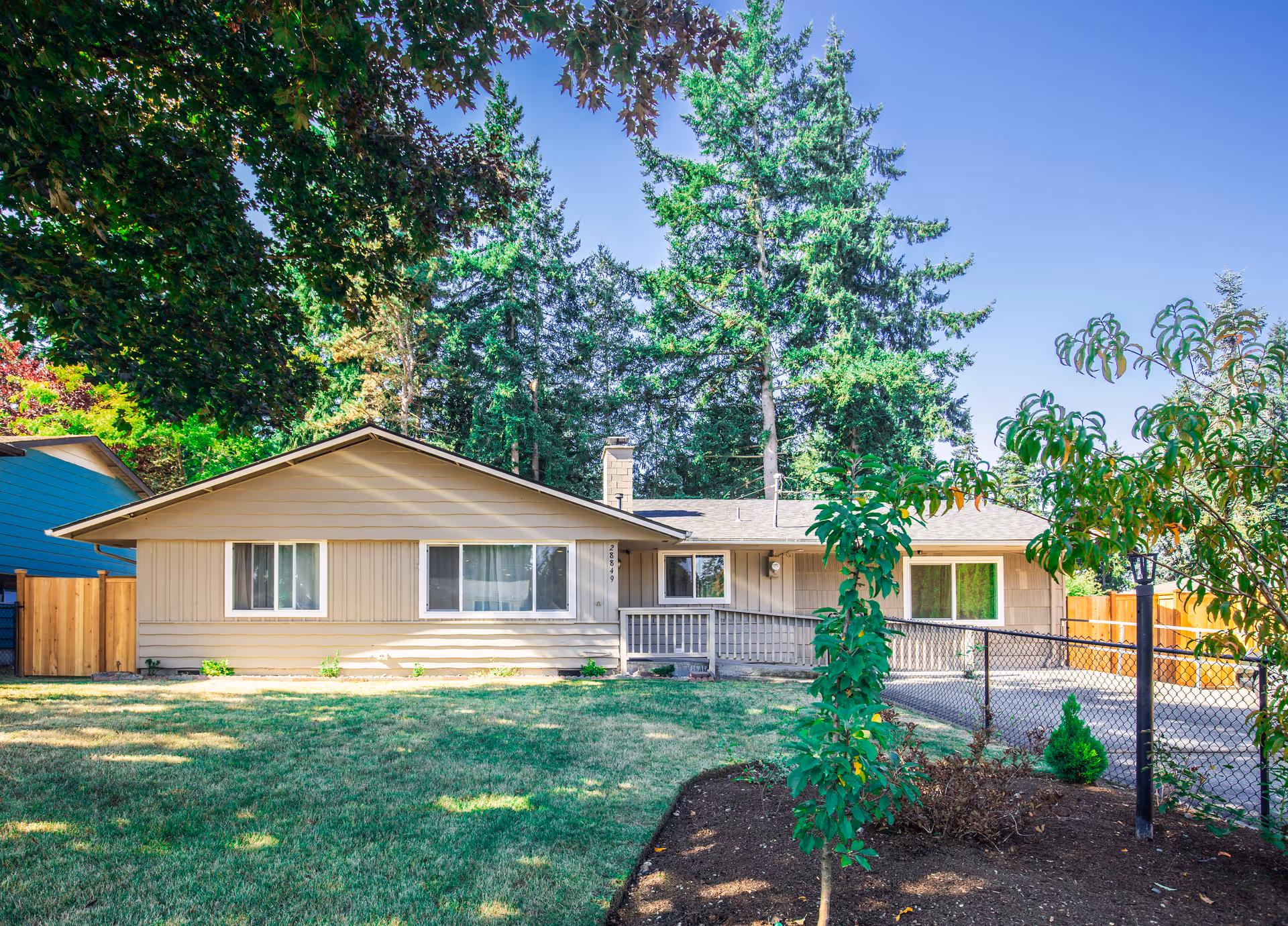 Single-story beige senior living building with front porch, lawn, and tall trees surrounding