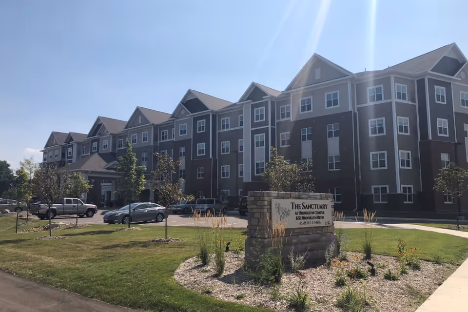 Exterior view of a multi-story senior living facility building with multiple windows, a parking lot with several cars, and a landscaped area with grass, small trees, and plants. A stone sign in the foreground reads 'The Sanctuary at Brooklyn Center, 6724 Brooklyn Blvd, Assisted Living.'