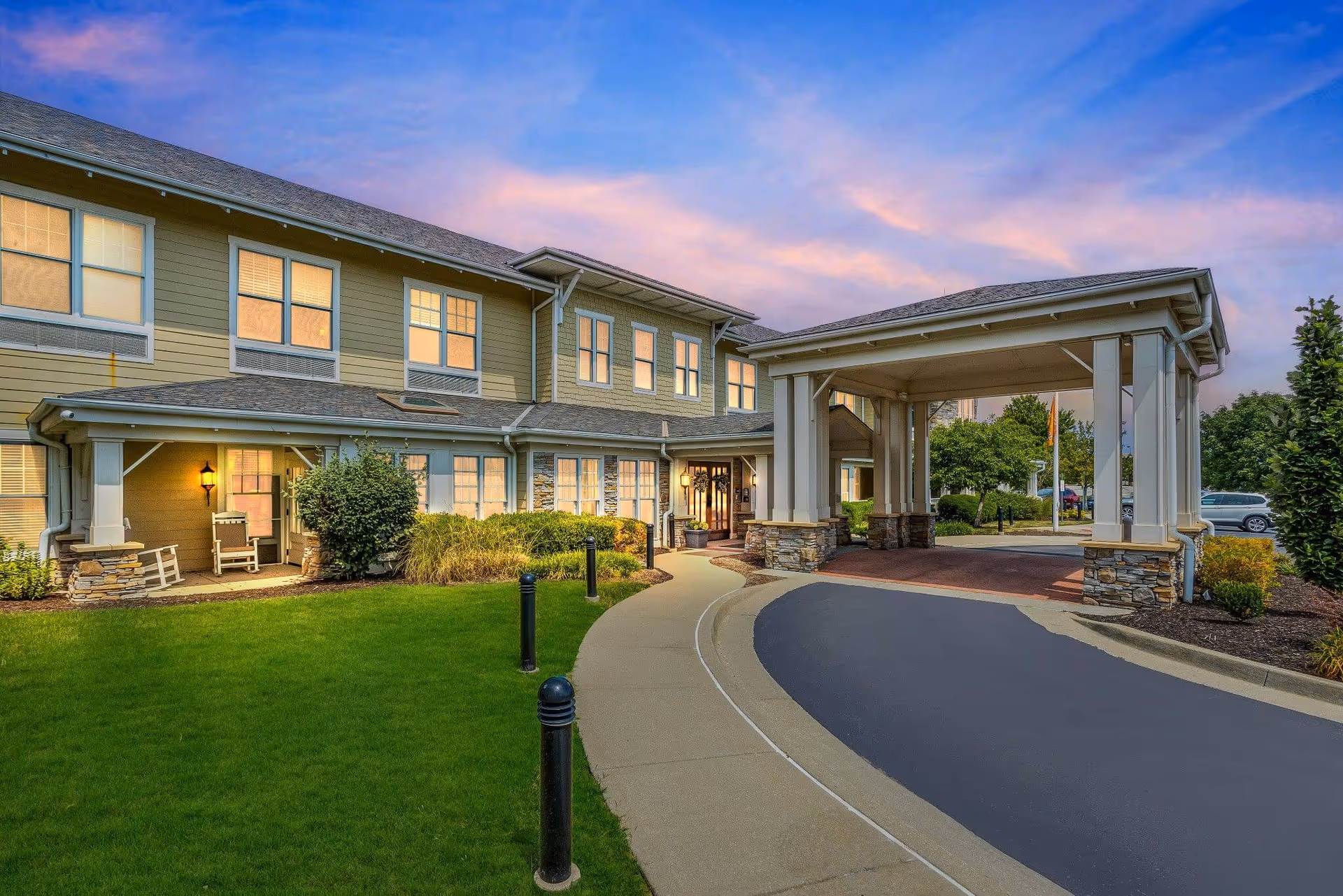Front entrance of a two-story senior living building with a covered porte-cochere, landscaped lawn, and illuminated windows at dusk.