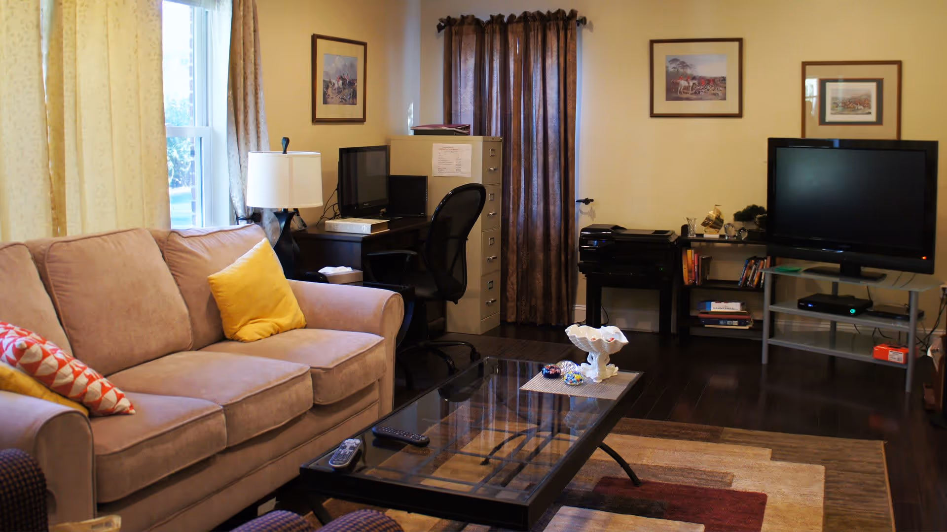 A cozy living room with a beige sofa adorned with colorful pillows, a glass coffee table with decorative items, a TV on a stand, a desk with a computer and office chair, a filing cabinet, and framed artwork on the walls. The room has wooden flooring and curtains on the windows.