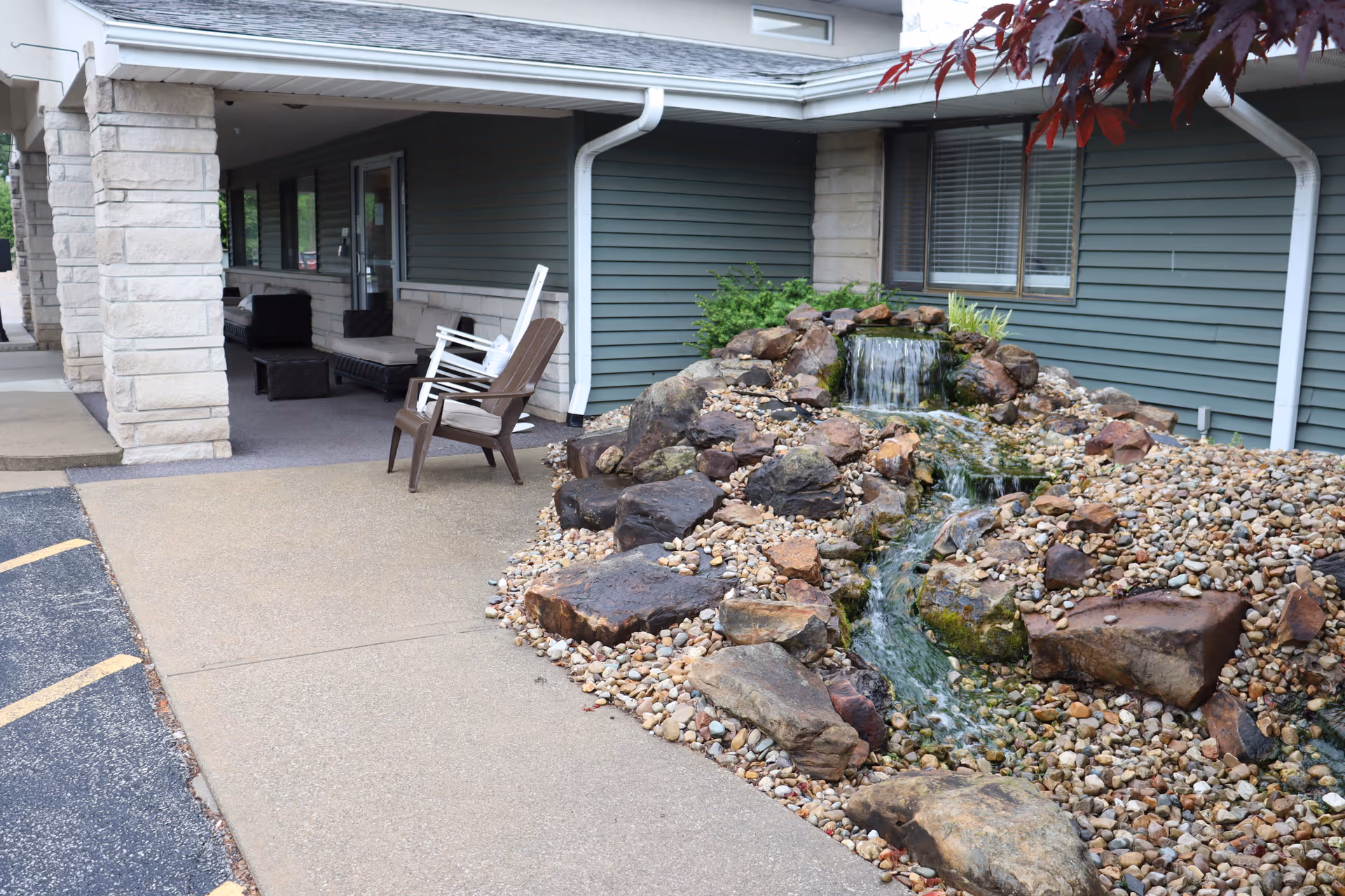 Covered entrance with stone columns, outdoor seating, and a rock waterfall feature beside the walkway.
