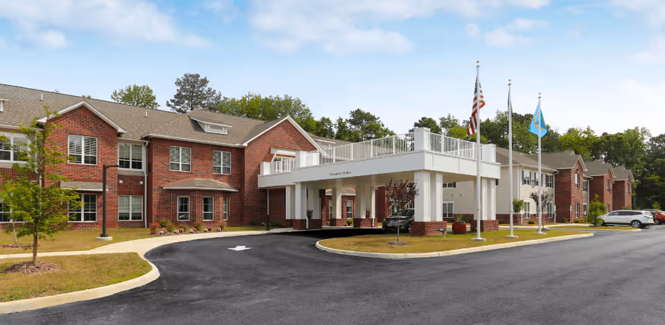 Exterior view of a two-story senior living facility with red brick and beige siding, a covered entrance with white pillars, three flagpoles with flags, a curved driveway, and several parked cars. The building is surrounded by green trees and a well-maintained lawn under a partly cloudy sky.