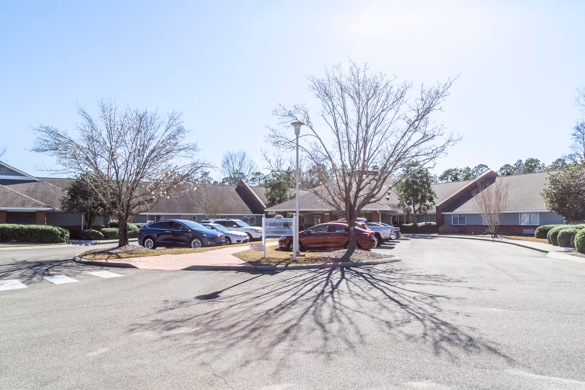 Exterior view of The Commons At Brightmore of Wilmington facility showing a parking area with several parked cars, leafless trees casting shadows on the pavement, and single-story buildings with sloped roofs in the background under a clear sky.
