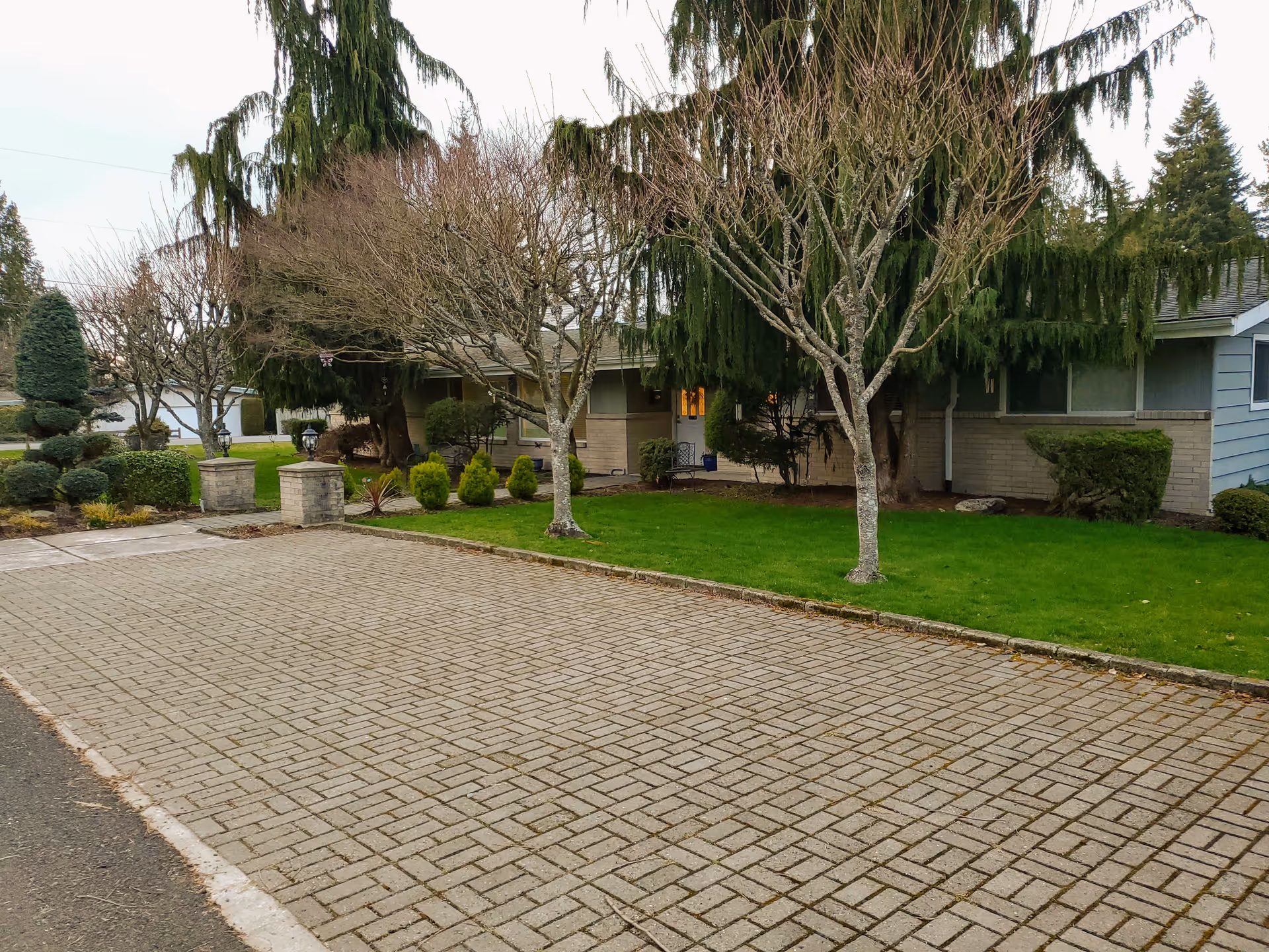 A paved driveway leading to a single-story building with a green lawn and several trees, including some with bare branches and others with dense green foliage. The building has a light-colored exterior with windows and a small porch area visible.