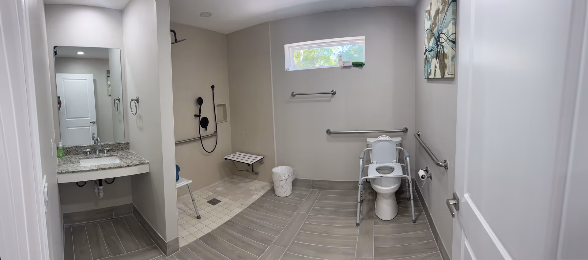 Accessible bathroom with sink, mirror, grab bars, shower area, toilet, and a window.