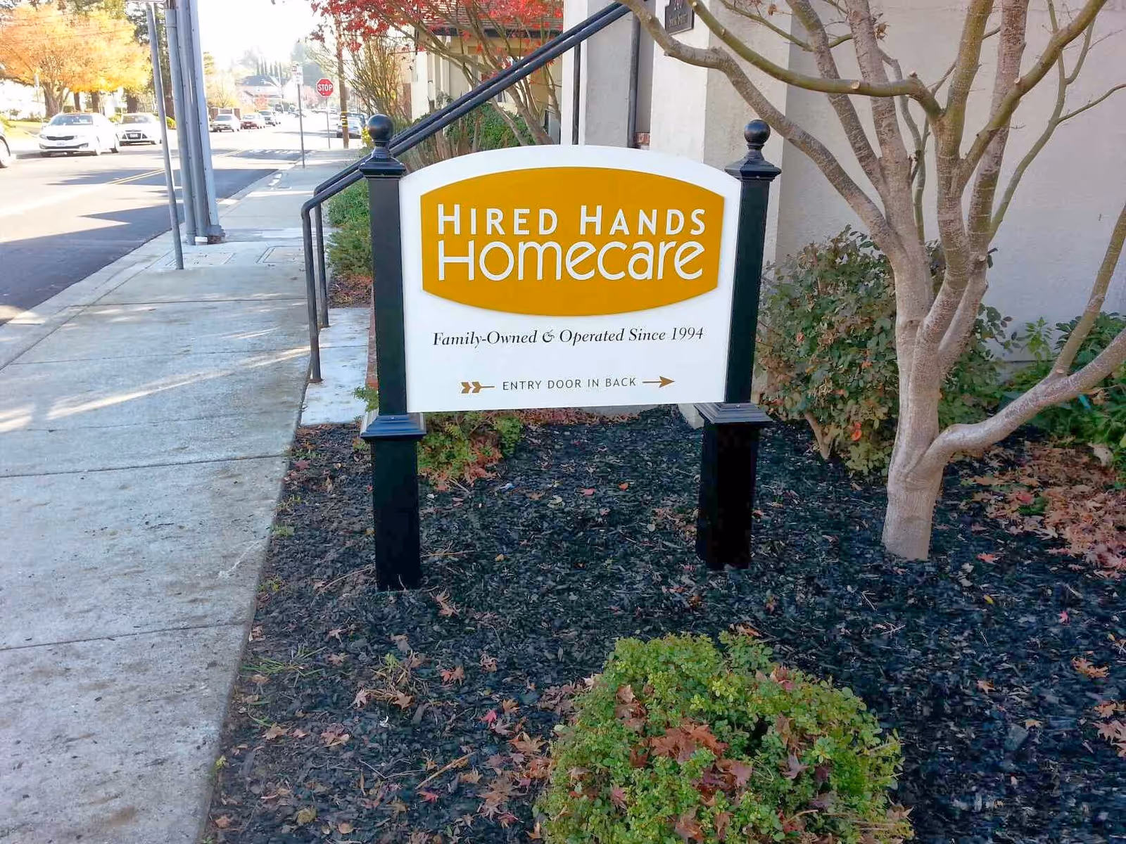 Exterior sidewalk with a Hired Hands Homecare Pleasanton sign beside trees and mulch.
