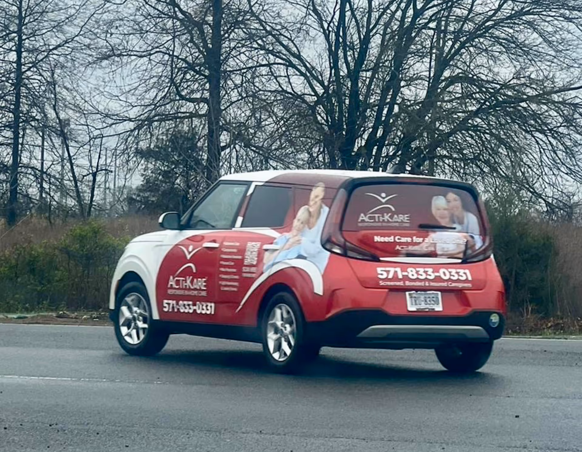 Red Acti-Kare van with photos and contact info parked on a road advertising senior care services.