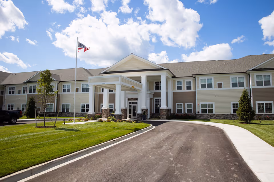Front exterior view of a two-story senior living facility building with a covered entrance supported by white columns. There is a neatly maintained lawn, a driveway leading to the entrance, an American flag on a flagpole, and a partly cloudy blue sky in the background.