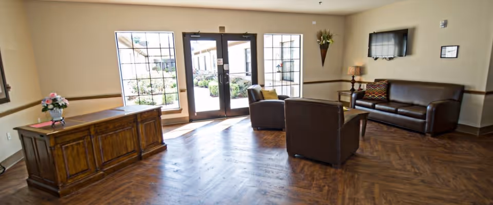 A bright and welcoming living room area with wooden flooring, a brown leather sofa, two matching armchairs, a wooden side table with a lamp, a wall-mounted TV, and a wooden desk with a flower vase. Large windows and glass doors let in natural light and provide a view of an outdoor garden area.