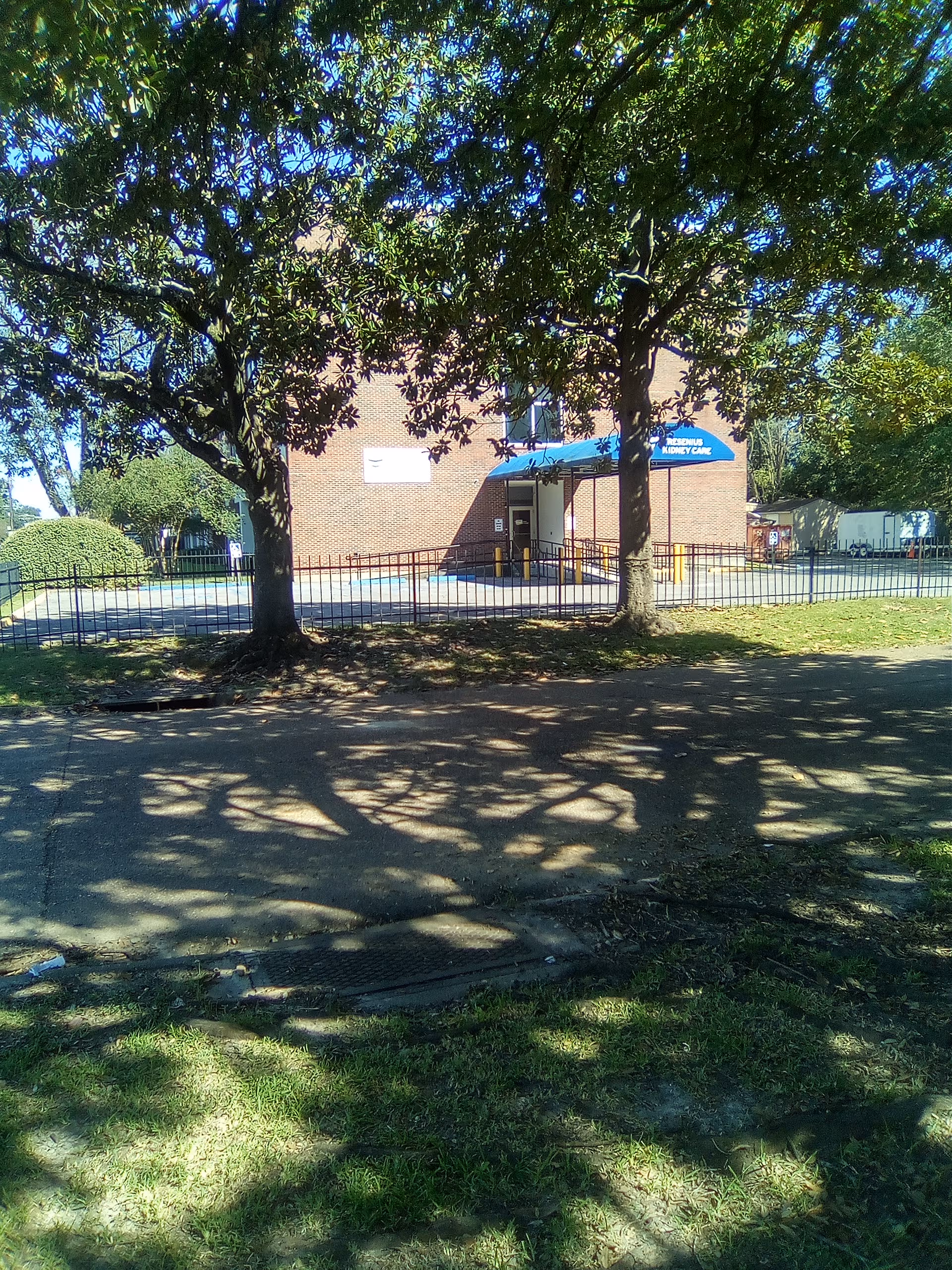 Brick building entrance with blue canopy, fenced walkway, trees casting shadows in foreground.