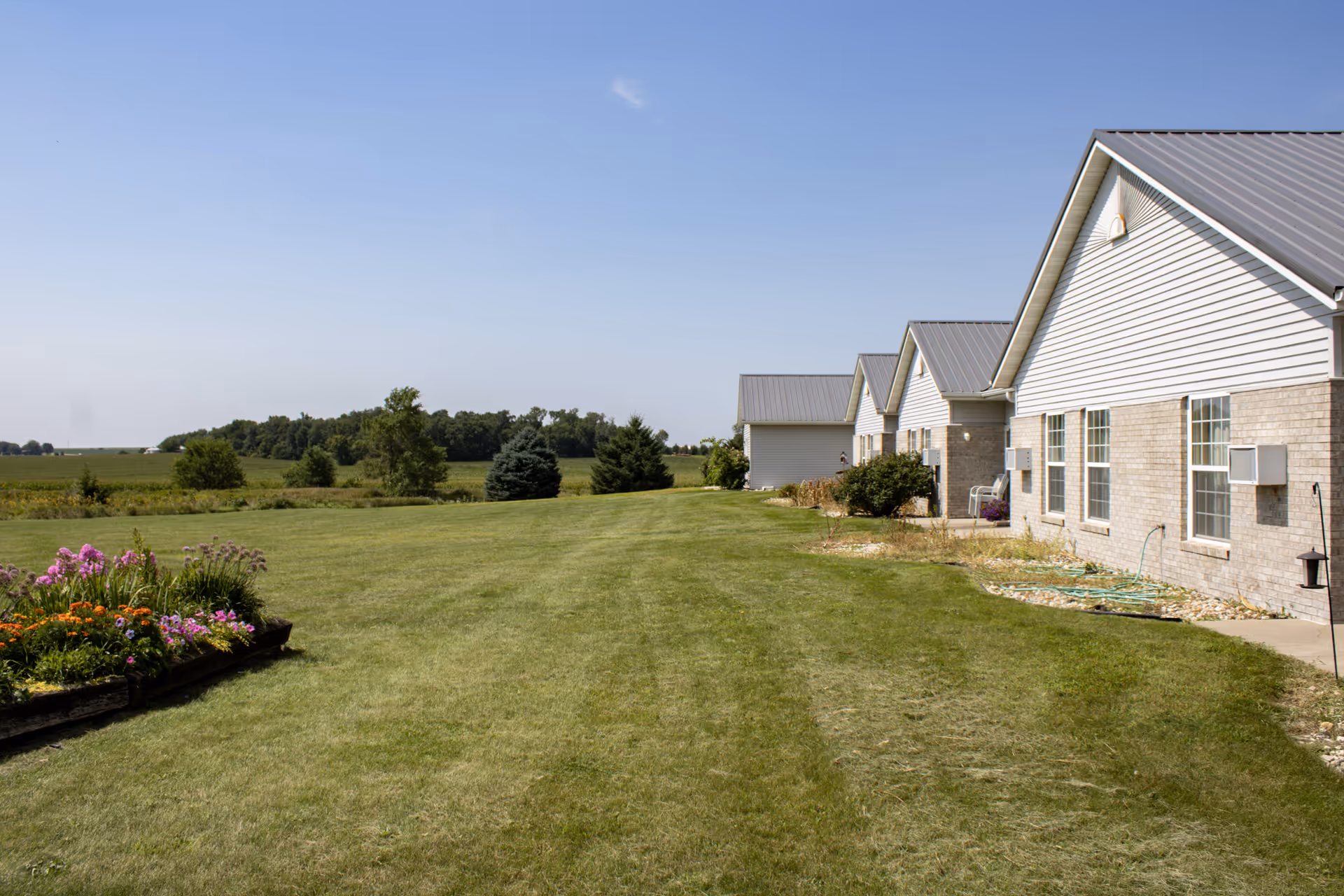 Row of light-brick senior living buildings with manicured lawn and flower beds under blue sky