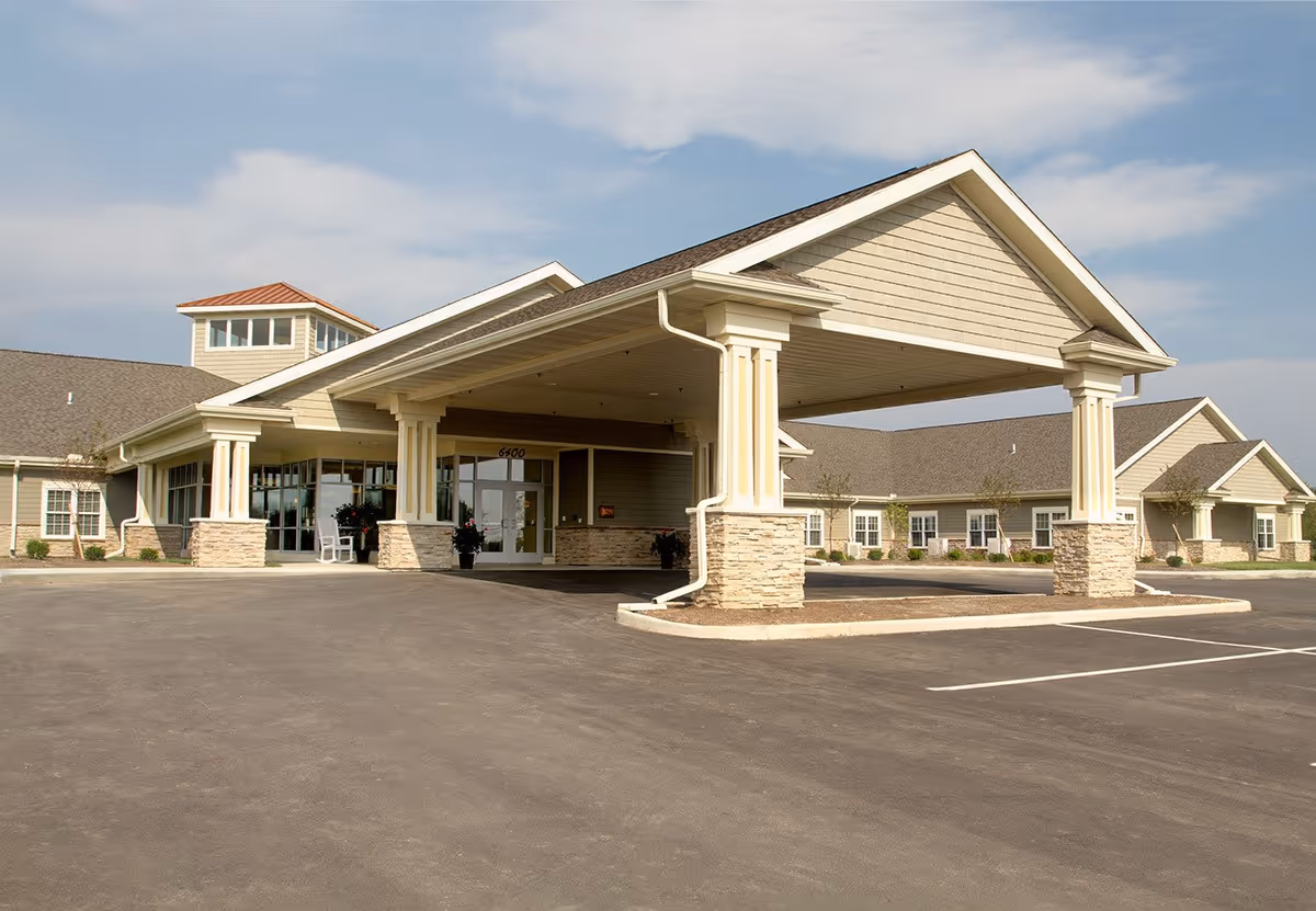 Exterior view of Randall Residence of Tipp City showing the front entrance with a large covered driveway supported by columns, surrounded by a paved parking area and landscaped with small bushes and trees under a partly cloudy sky.