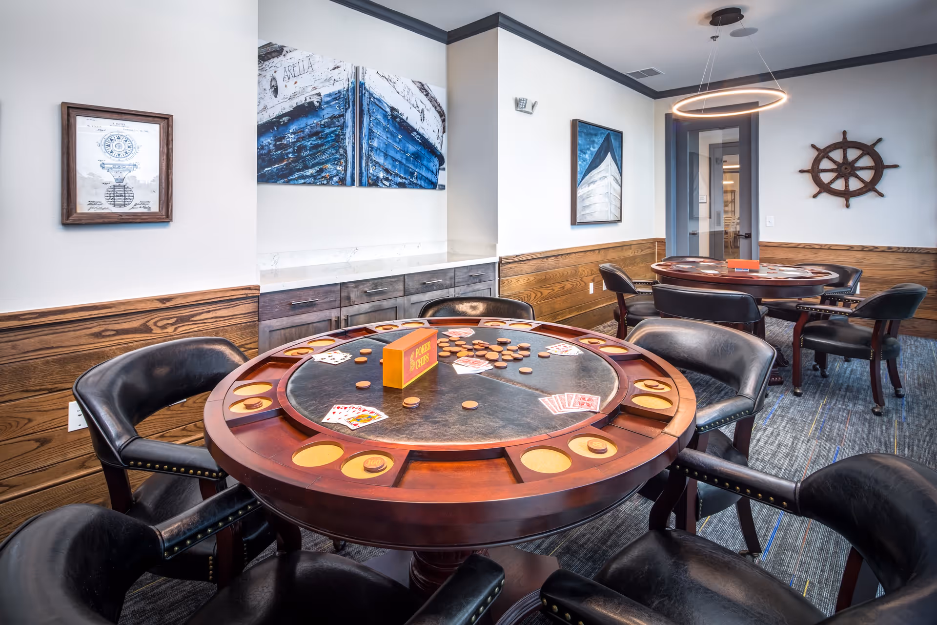 Interior view of a game room in Arella on Jones Active Senior Community featuring two round wooden game tables with black leather chairs around them. The tables have poker chips, playing cards, and a sign that says 'Poker Chips'. The room has wood paneling on the lower half of the walls, framed nautical-themed artwork, a ship's wheel mounted on the wall, and a modern circular light fixture hanging from the ceiling.