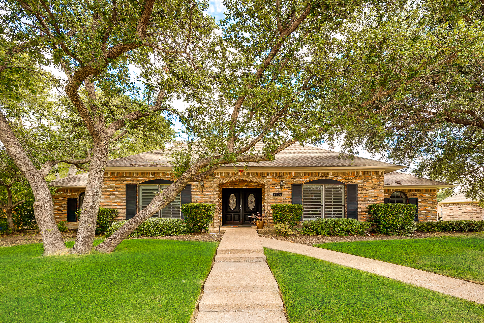 Front exterior view of a single-story brick building with a walkway leading to a double-door entrance. The building is surrounded by green grass, bushes, and large trees with sprawling branches.